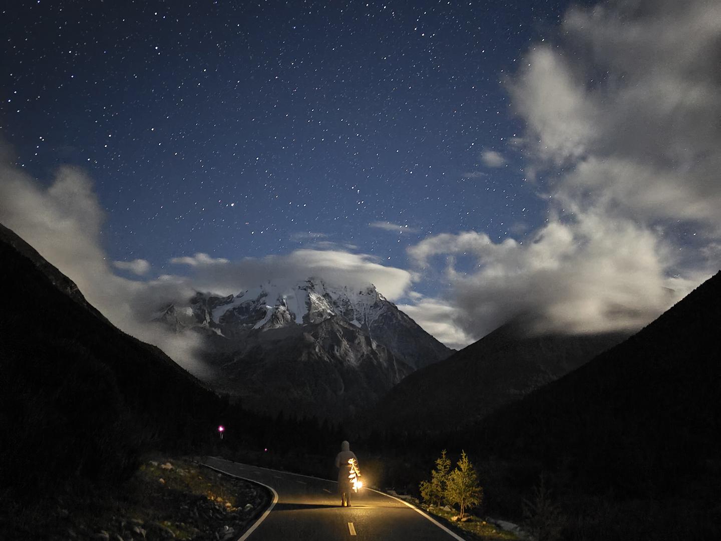 A lone man stands on a winding mountain road under a star-filled night sky with snow-capped peaks in the background, captured with stunning clarity and contrast.