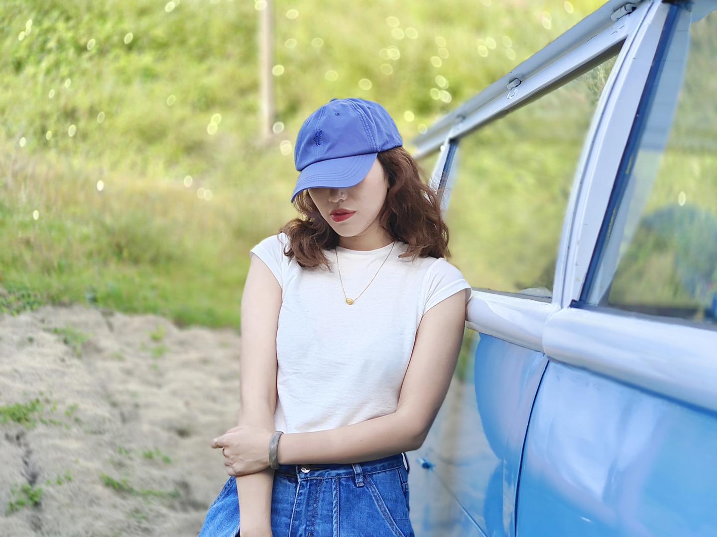A woman wearing a blue cap and white shirt stands beside a blue vehicle in a grassy outdoor setting, captured in sharp detail with a ZEISS lens.