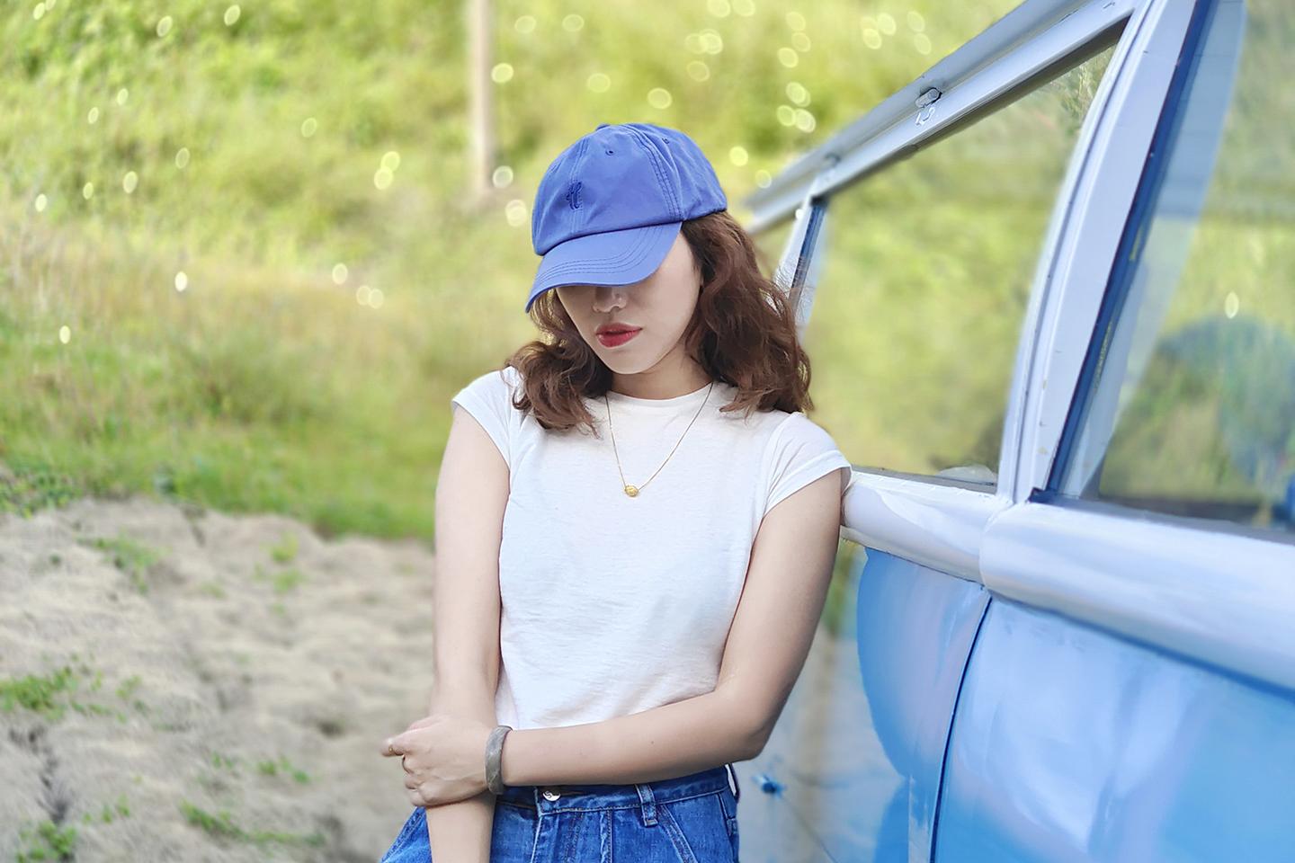 A woman wearing a blue cap and white shirt stands beside a blue vehicle in a grassy outdoor setting, captured in sharp detail with a ZEISS lens.