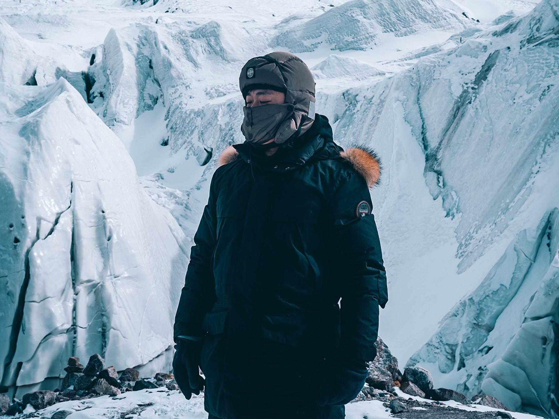 A person dressed in heavy winter gear stands on a snowy, icy mountainside surrounded by large glaciers.