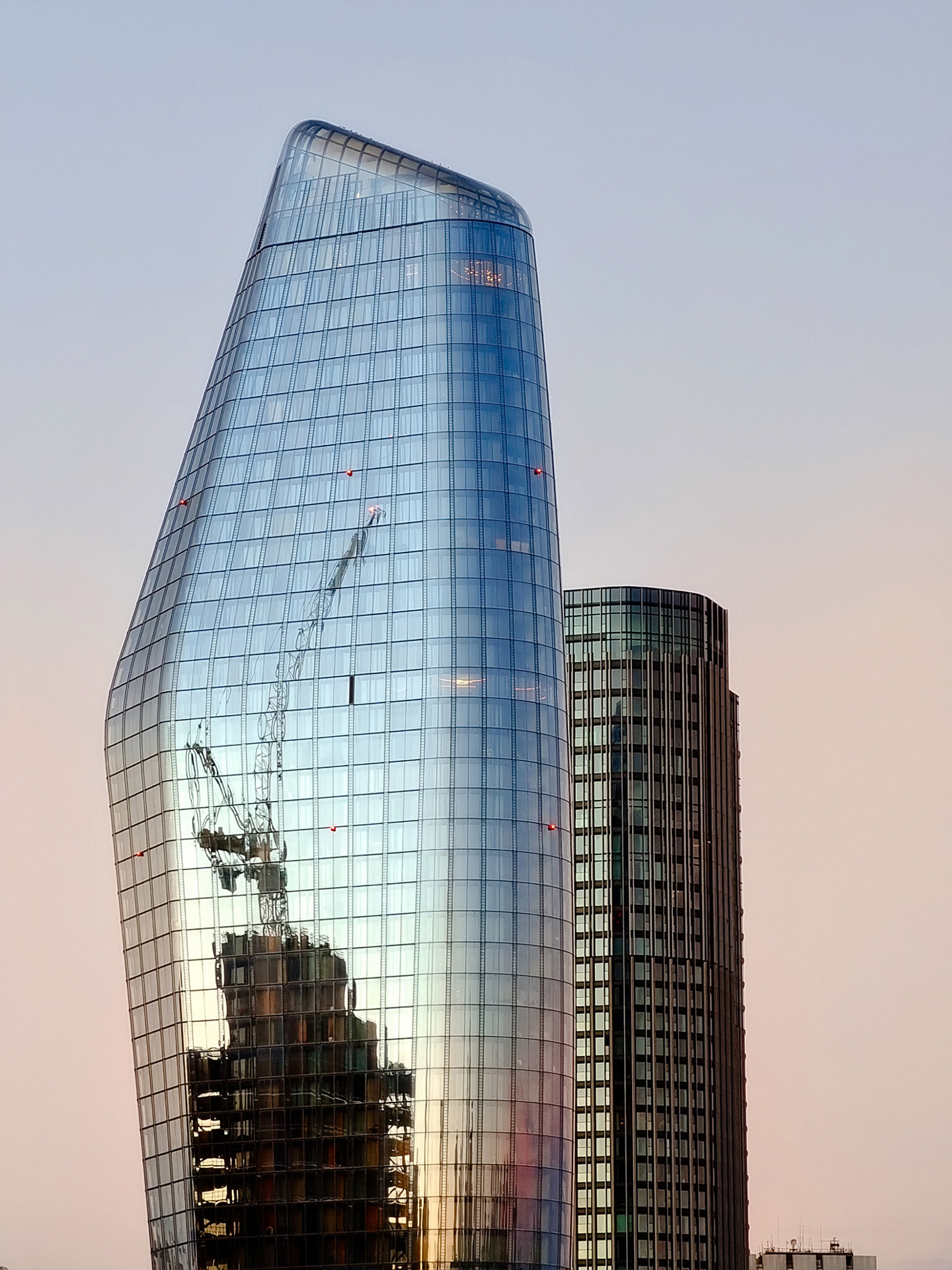 Modern glass skyscraper reflecting a construction site with cranes during sunset, adjacent to a cylindrical office building.