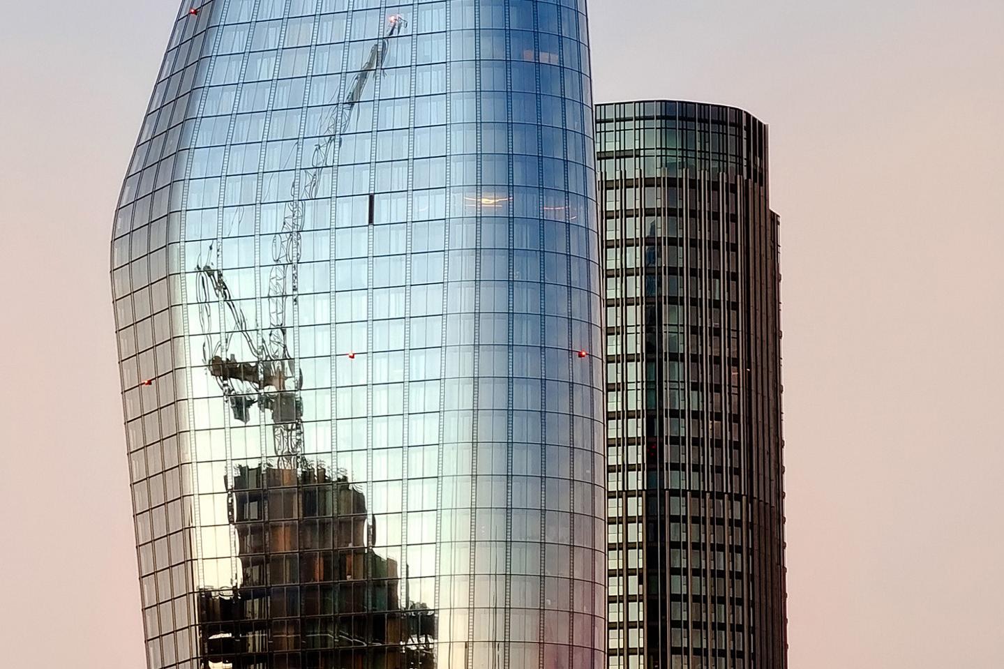 Modern glass skyscraper reflecting a construction site with cranes during sunset, adjacent to a cylindrical office building.