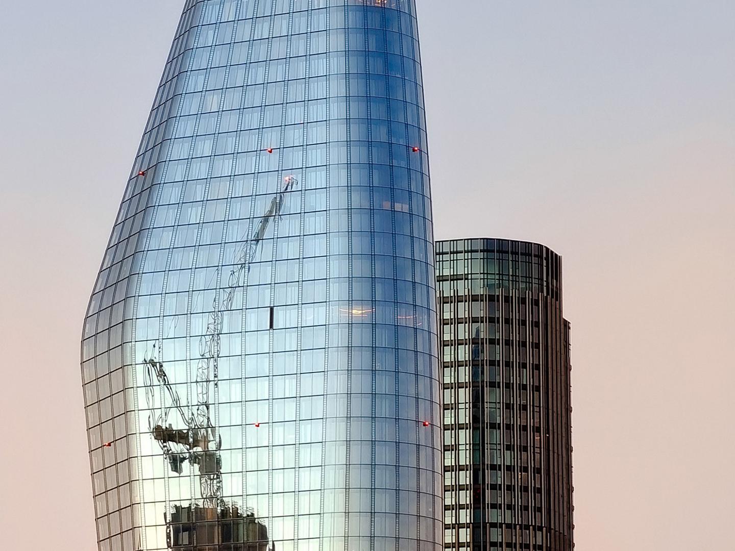 Modern glass skyscraper reflecting a construction site with cranes during sunset, adjacent to a cylindrical office building.
