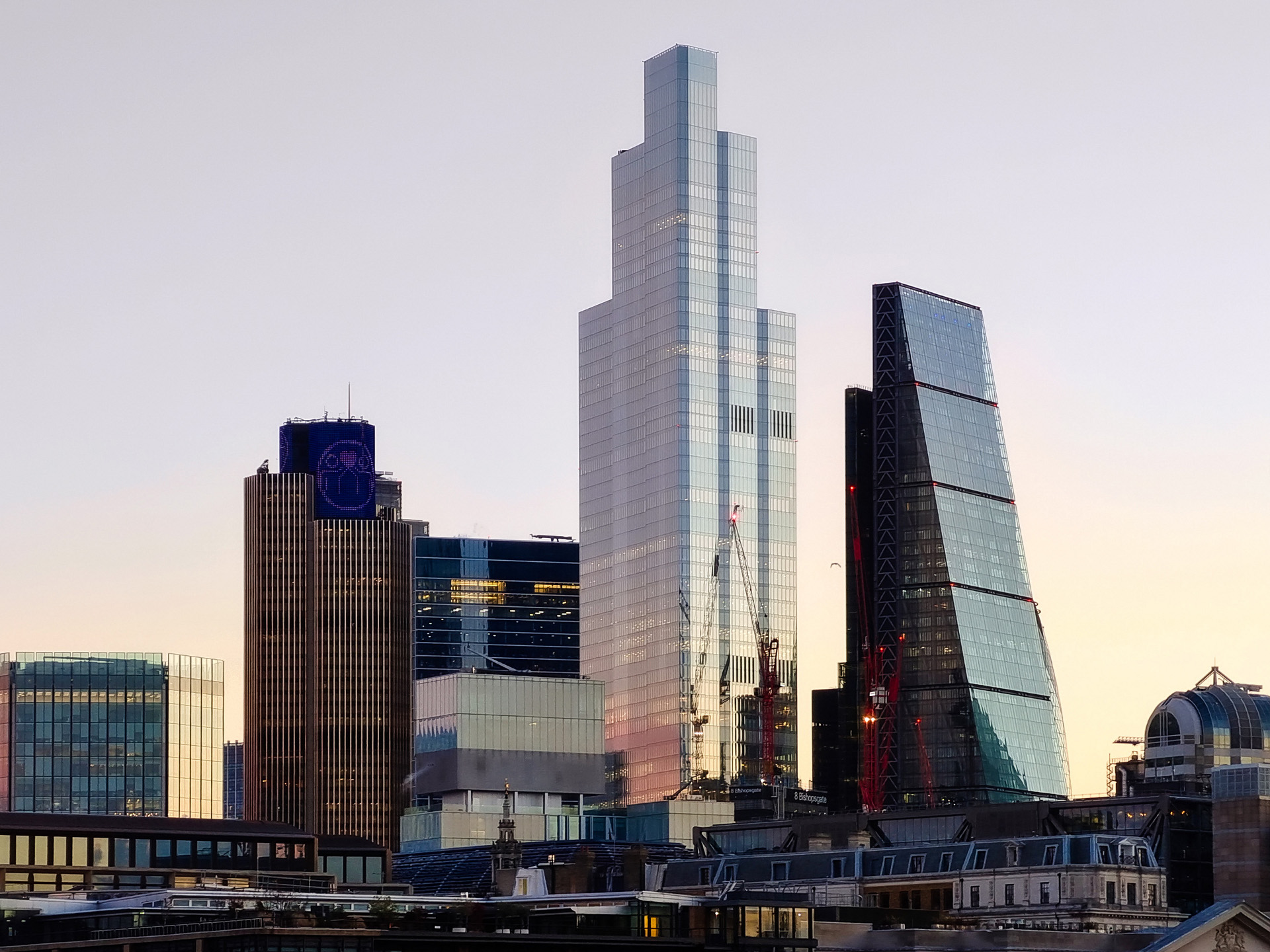 Modern city skyline at sunset featuring a cluster of high-rise office buildings with cranes, reflecting glass facades, and diverse architectural styles.
