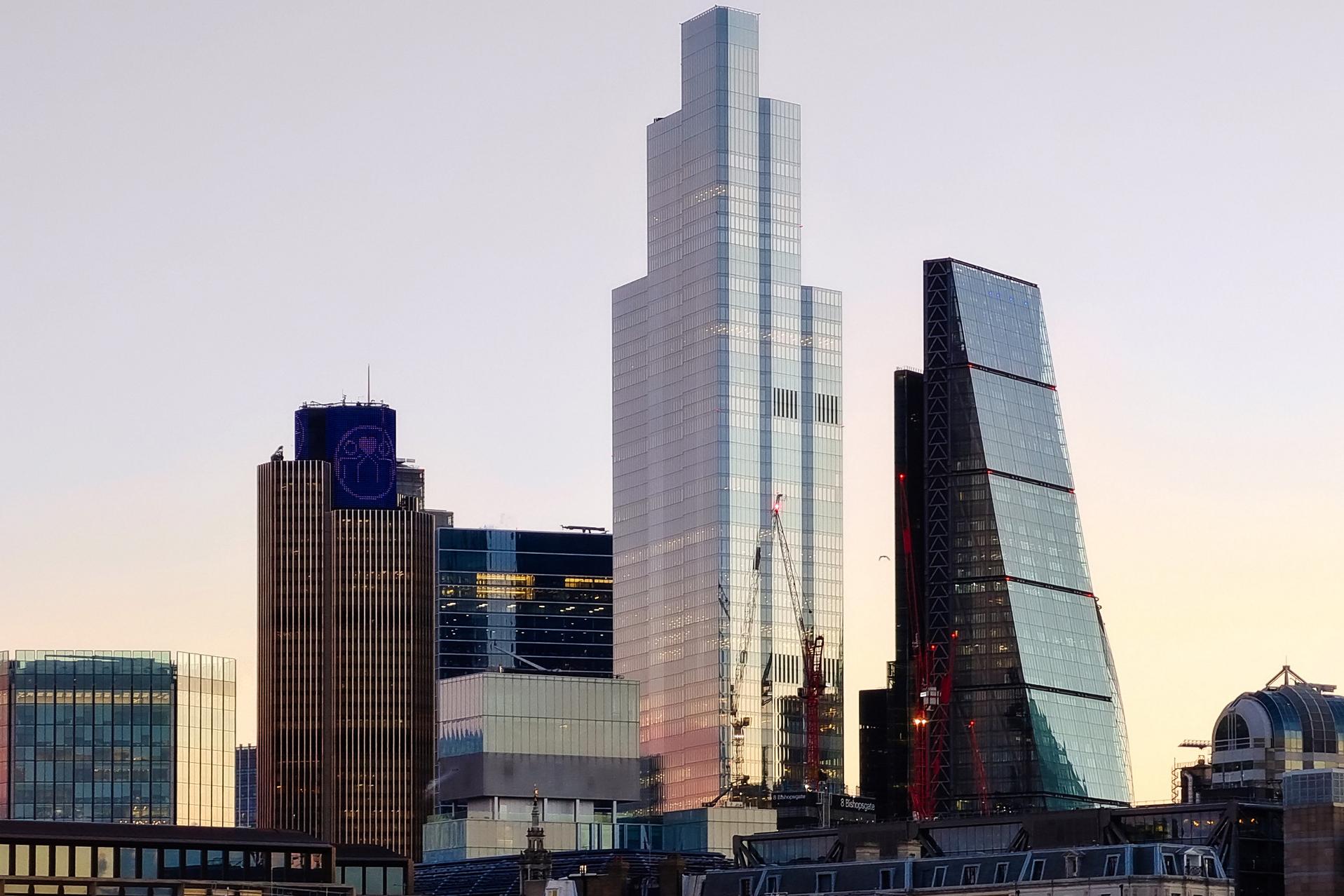Modern city skyline at sunset featuring a cluster of high-rise office buildings with cranes, reflecting glass facades, and diverse architectural styles.