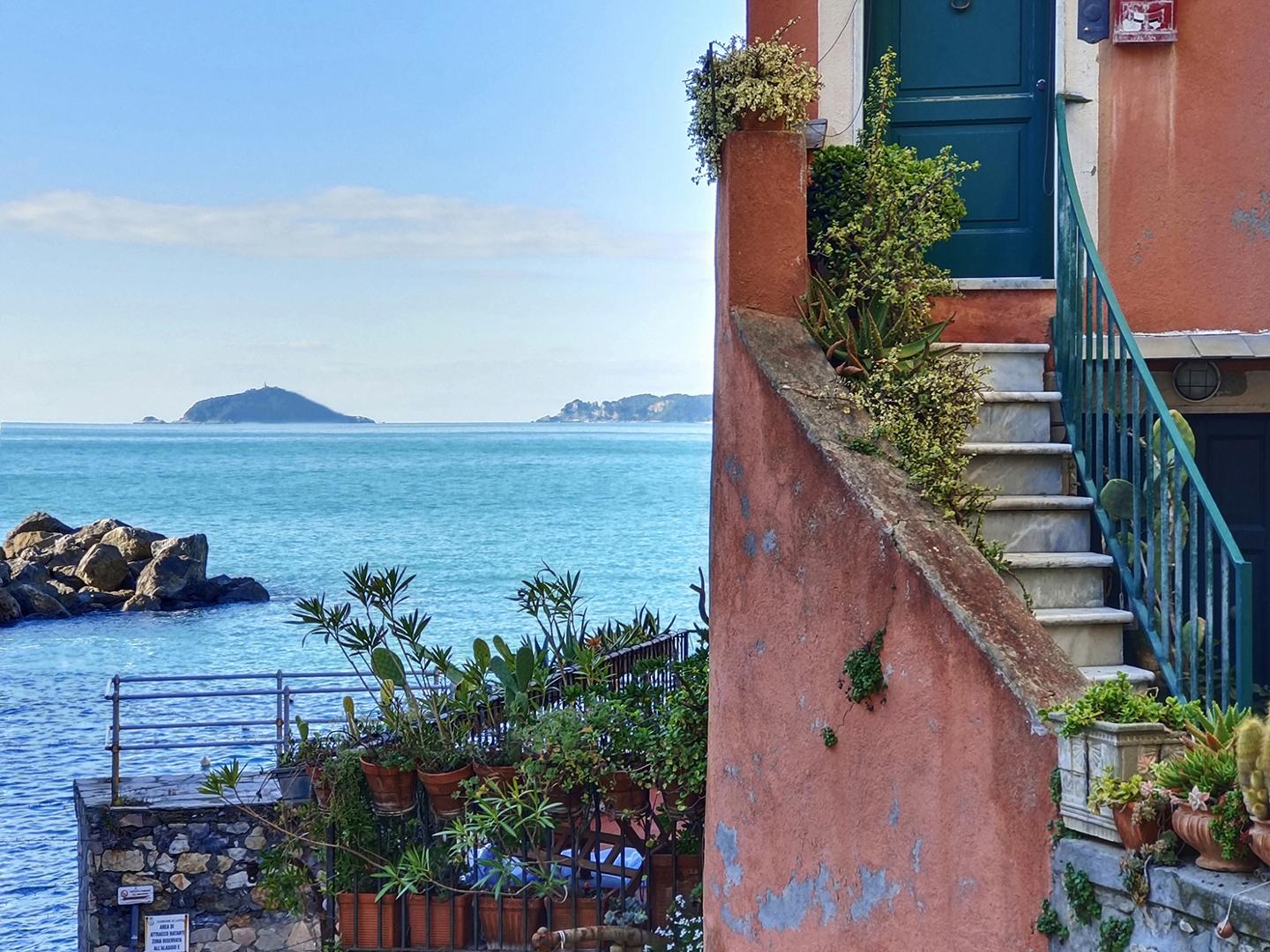 A seaside house with a terracotta exterior, stone steps, and potted plants overlooks a calm blue ocean under a clear sky, captured in vivid detail by ZEISS optics.