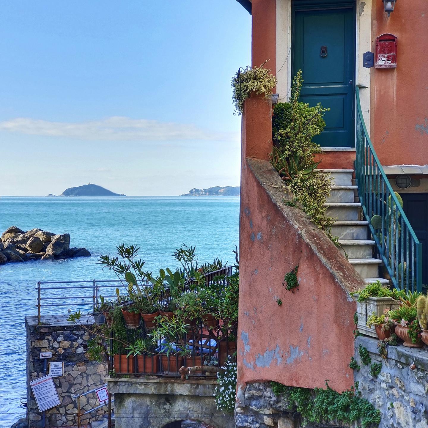 A seaside house with a terracotta exterior, stone steps, and potted plants overlooks a calm blue ocean under a clear sky, captured in vivid detail by ZEISS optics.