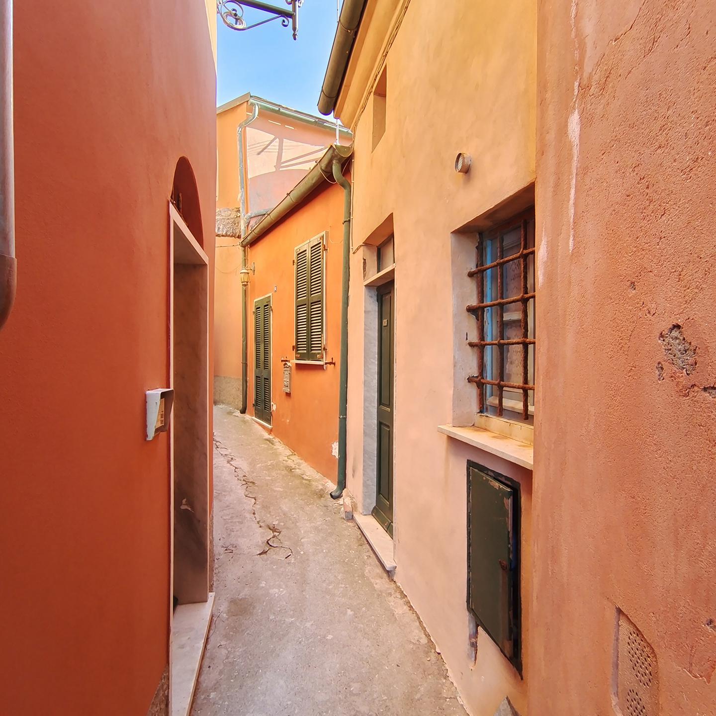 Narrow alleyway between colorful, sunlit buildings with textured walls and shuttered windows, captured in sharp detail with ZEISS optics.