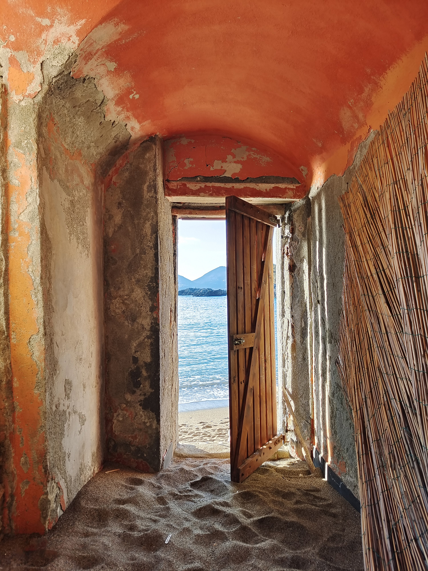 A rustic doorway with a partially open wooden door reveals a serene beach and calm sea with mountains in the distance, viewed from inside a weathered, sand-covered room.