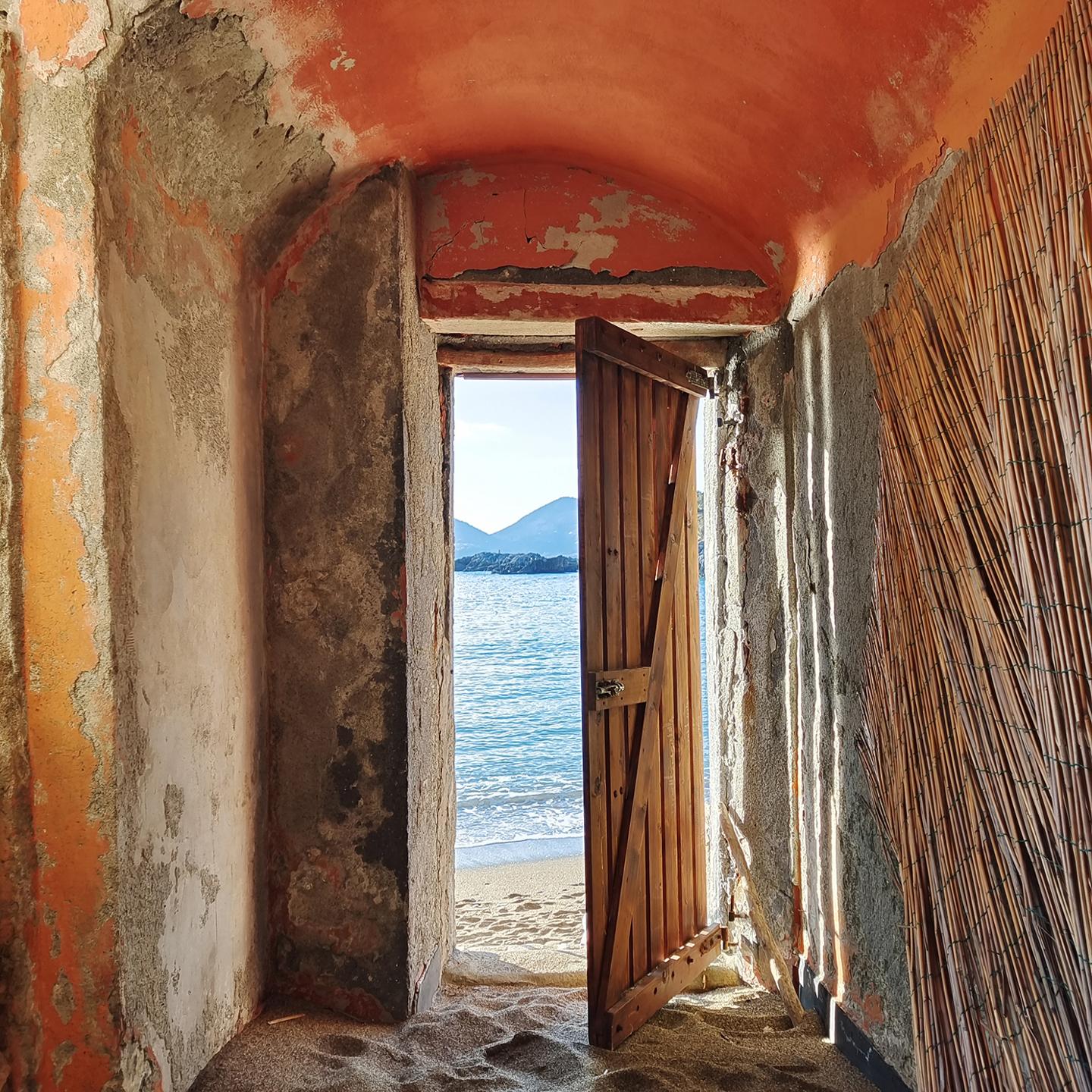A rustic doorway with a partially open wooden door reveals a serene beach and calm sea with mountains in the distance, viewed from inside a weathered, sand-covered room.