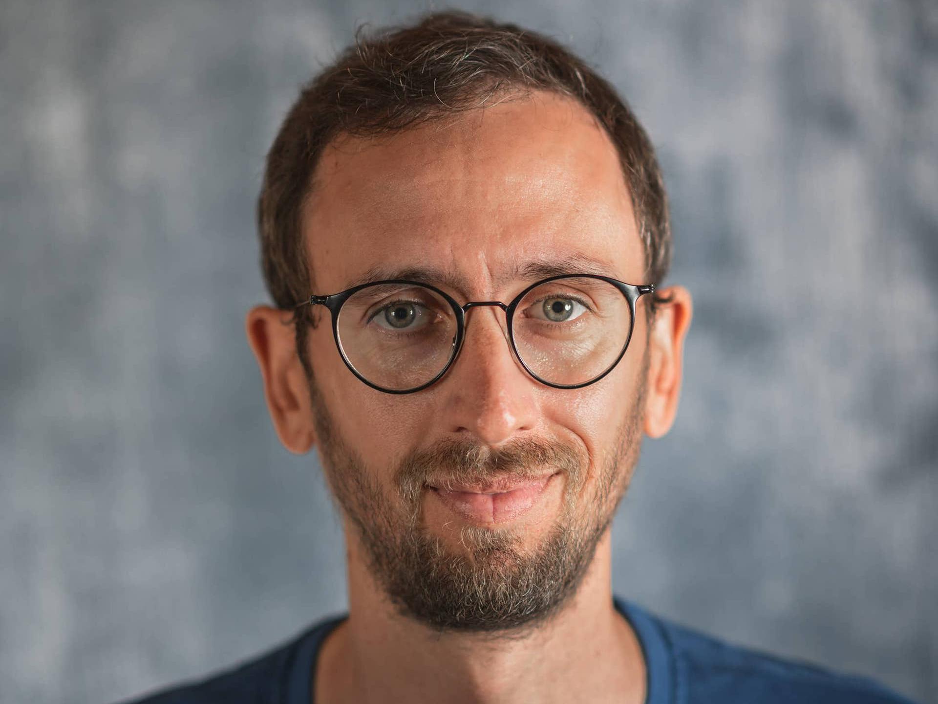 Man wearing round ZEISS eyeglass lenses and a blue shirt, standing in front of a neutral textured background.