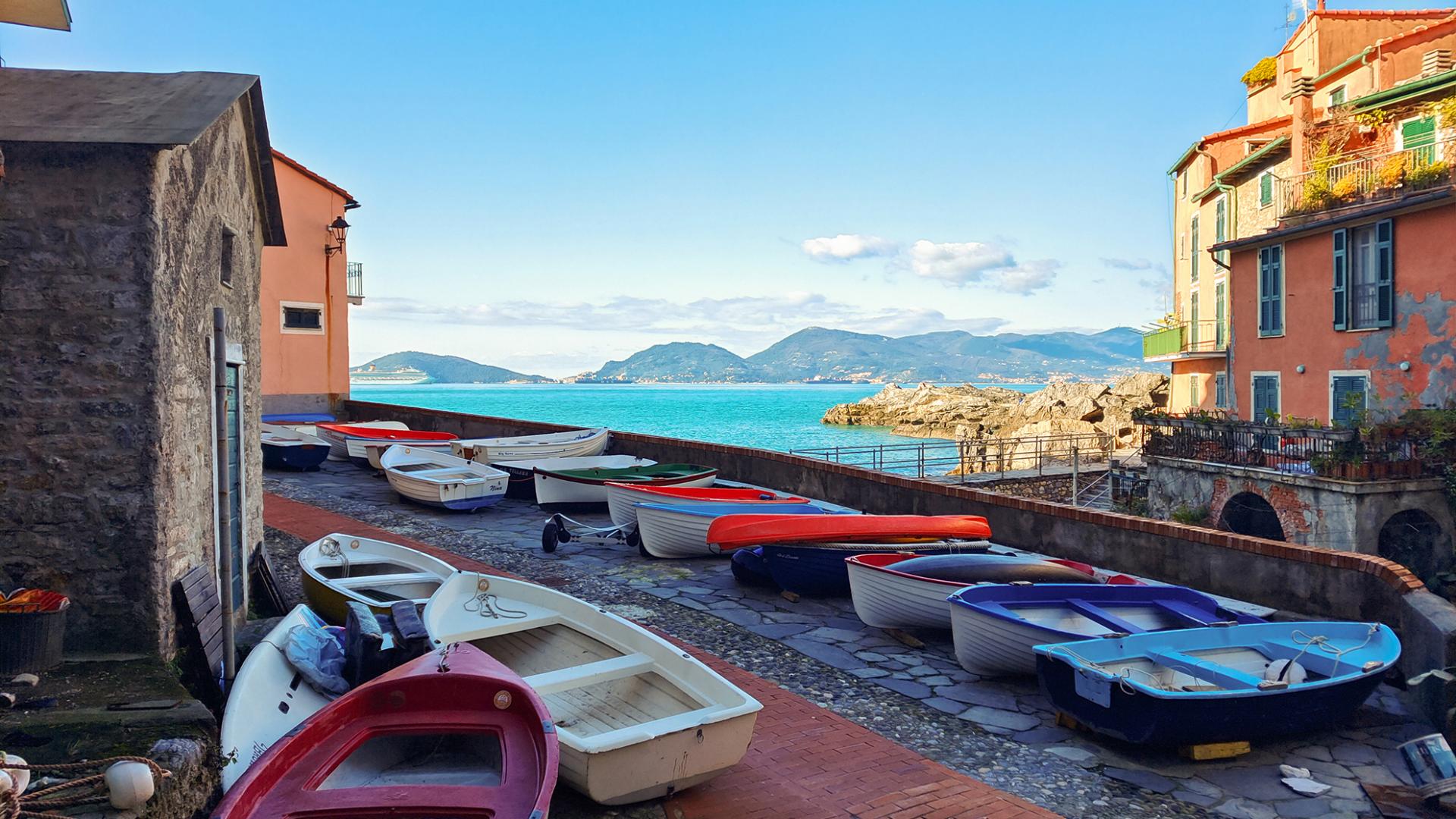 Colorful rowboats are lined up along a stone walkway in a coastal village with a scenic view of the sea and distant mountains, captured with ZEISS clarity.