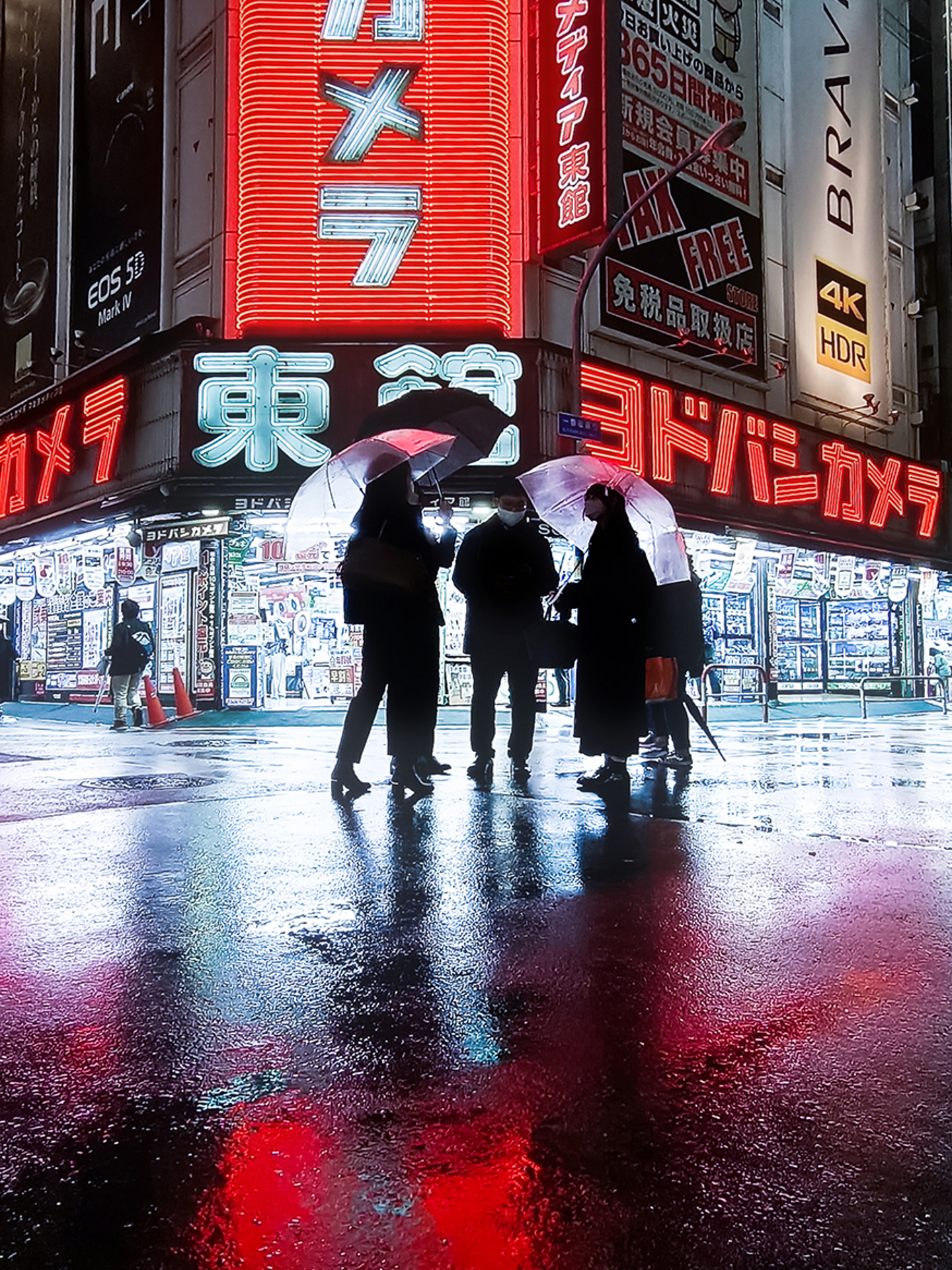 Four people holding transparent umbrellas stand on a wet, reflective street in front of brightly lit electronics stores at night.
