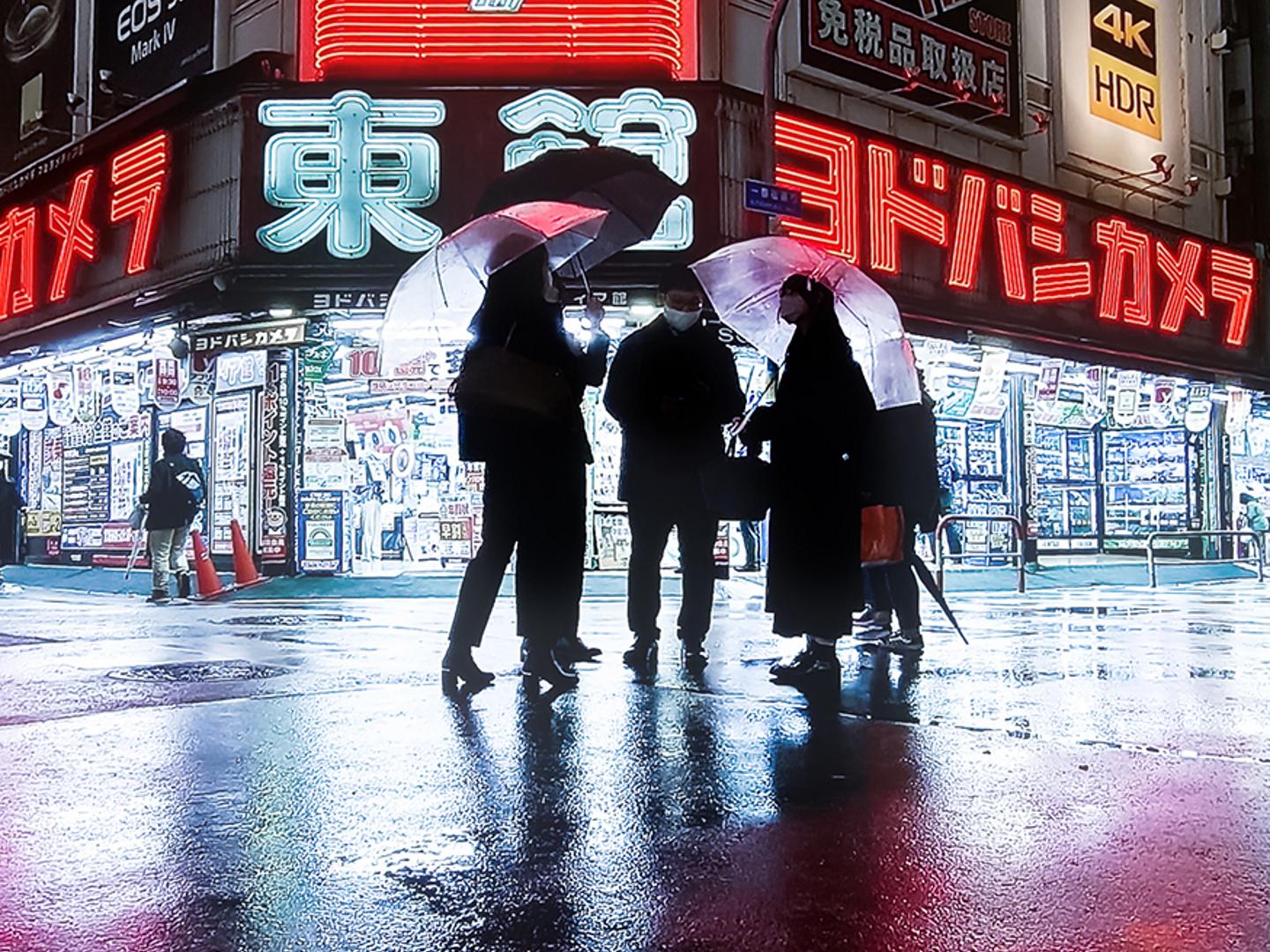 Four people holding transparent umbrellas stand on a wet, reflective street in front of brightly lit electronics stores at night.