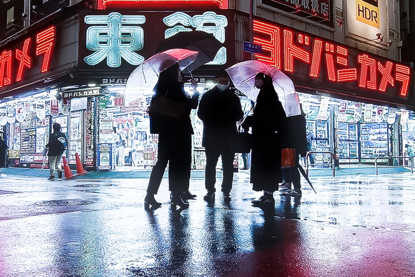 Four people holding transparent umbrellas stand on a wet, reflective street in front of brightly lit electronics stores at night.