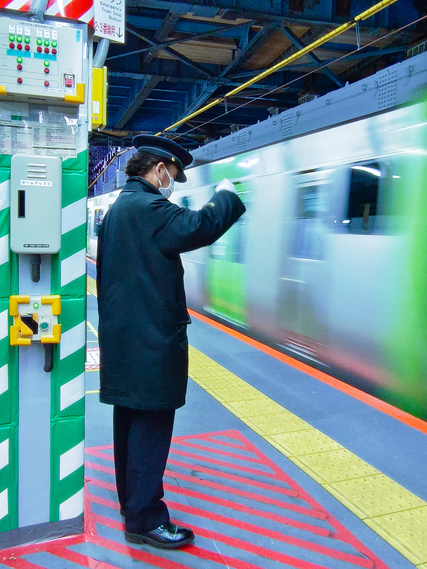 A train station worker in uniform and face mask signals beside a passing train on a brightly lit platform.