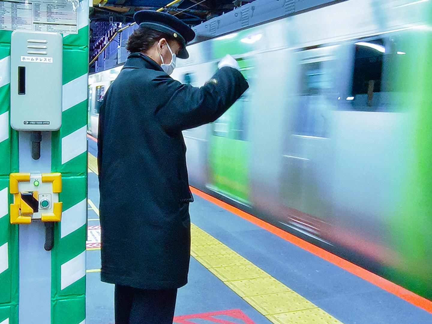 A train station worker in uniform and face mask signals beside a passing train on a brightly lit platform.