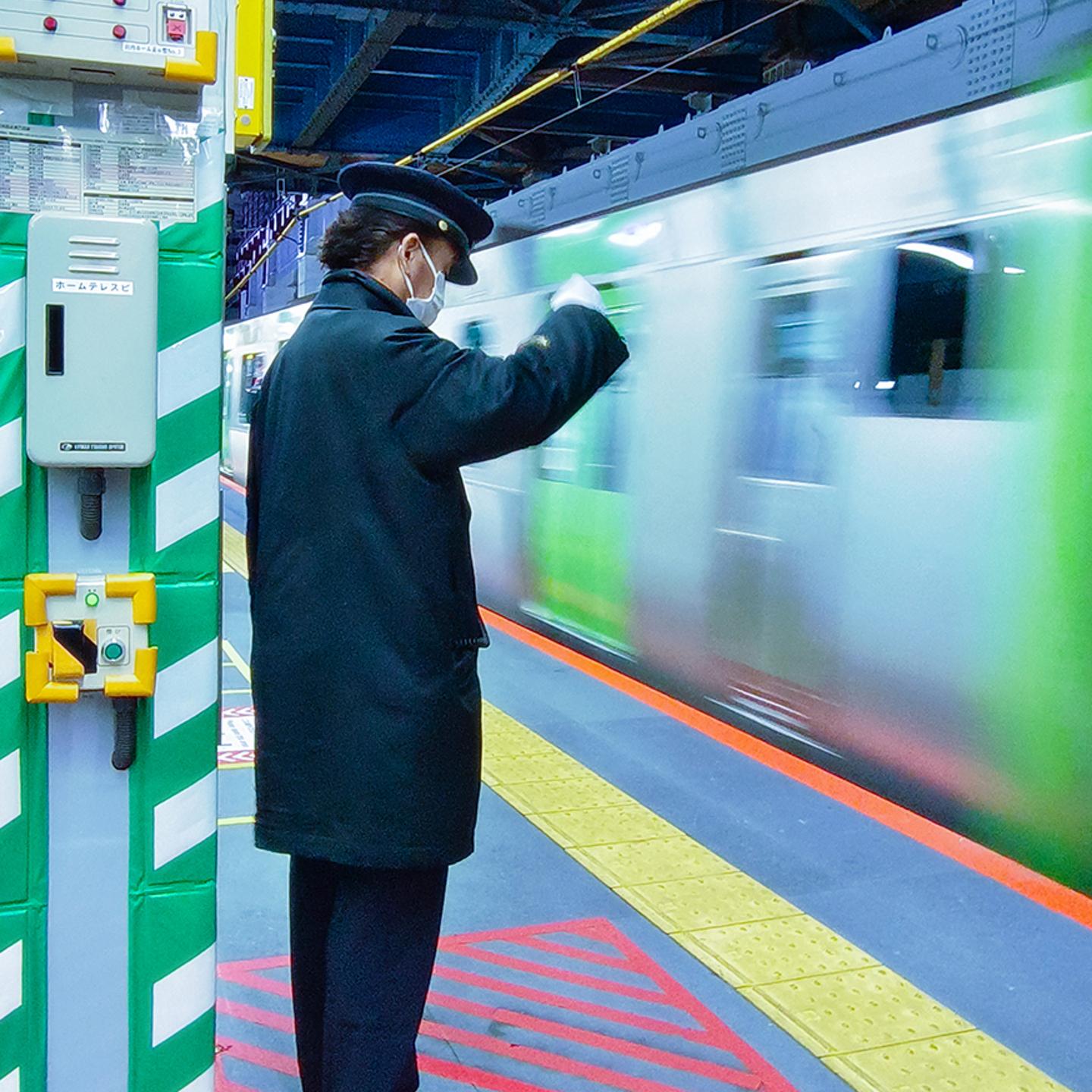 A train station worker in uniform and face mask signals beside a passing train on a brightly lit platform.