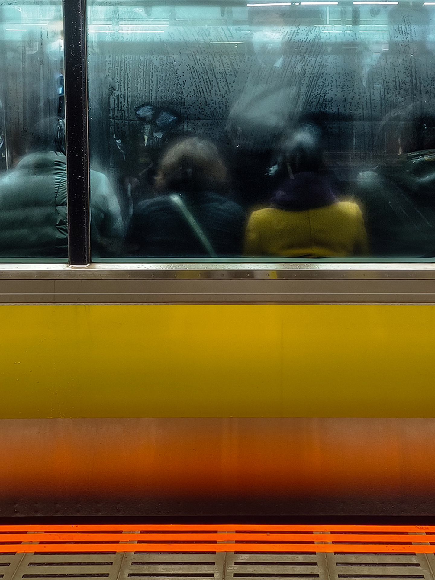 Passengers are seen through a fogged and reflective window of a yellow and orange subway train.