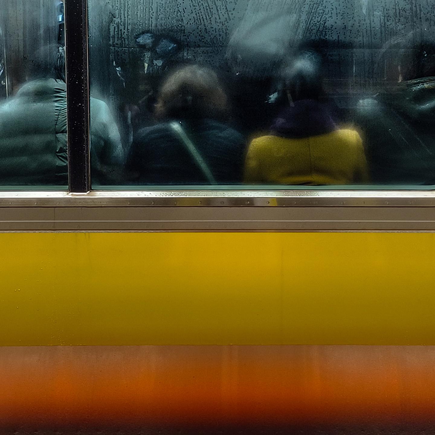 Passengers are seen through a fogged and reflective window of a yellow and orange subway train.