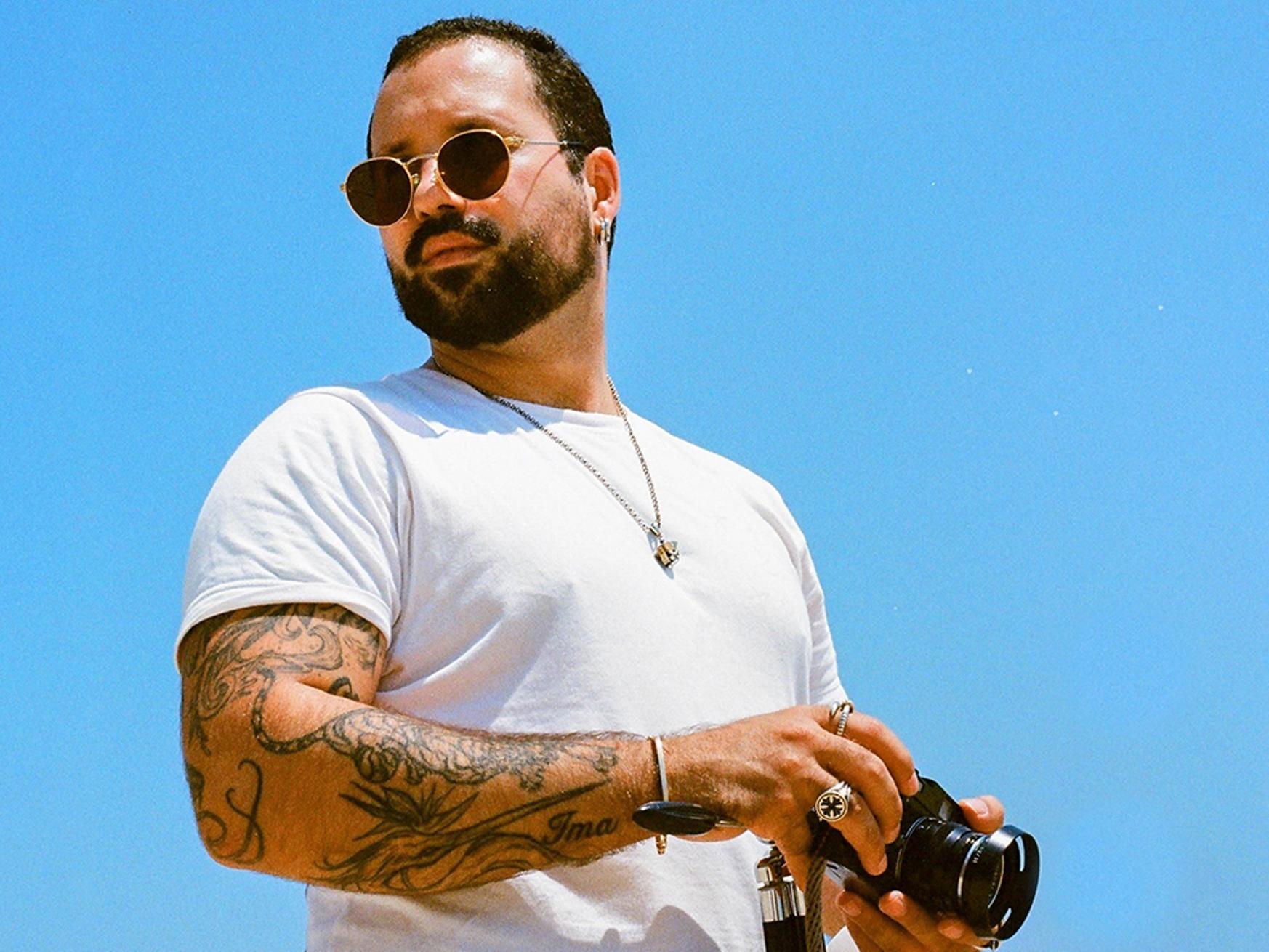 Man wearing sunglasses and a white t-shirt holding a camera under a clear blue sky, with visible ZEISS lens.