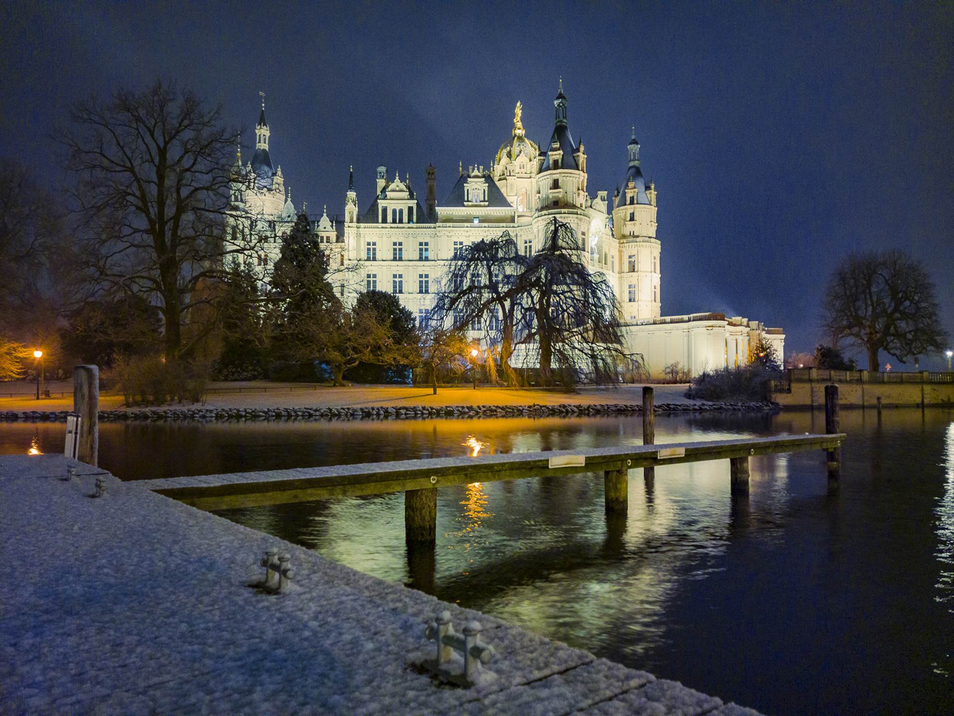 Illuminated historic castle at night reflected in calm water, viewed from a snowy dock under a dark sky.