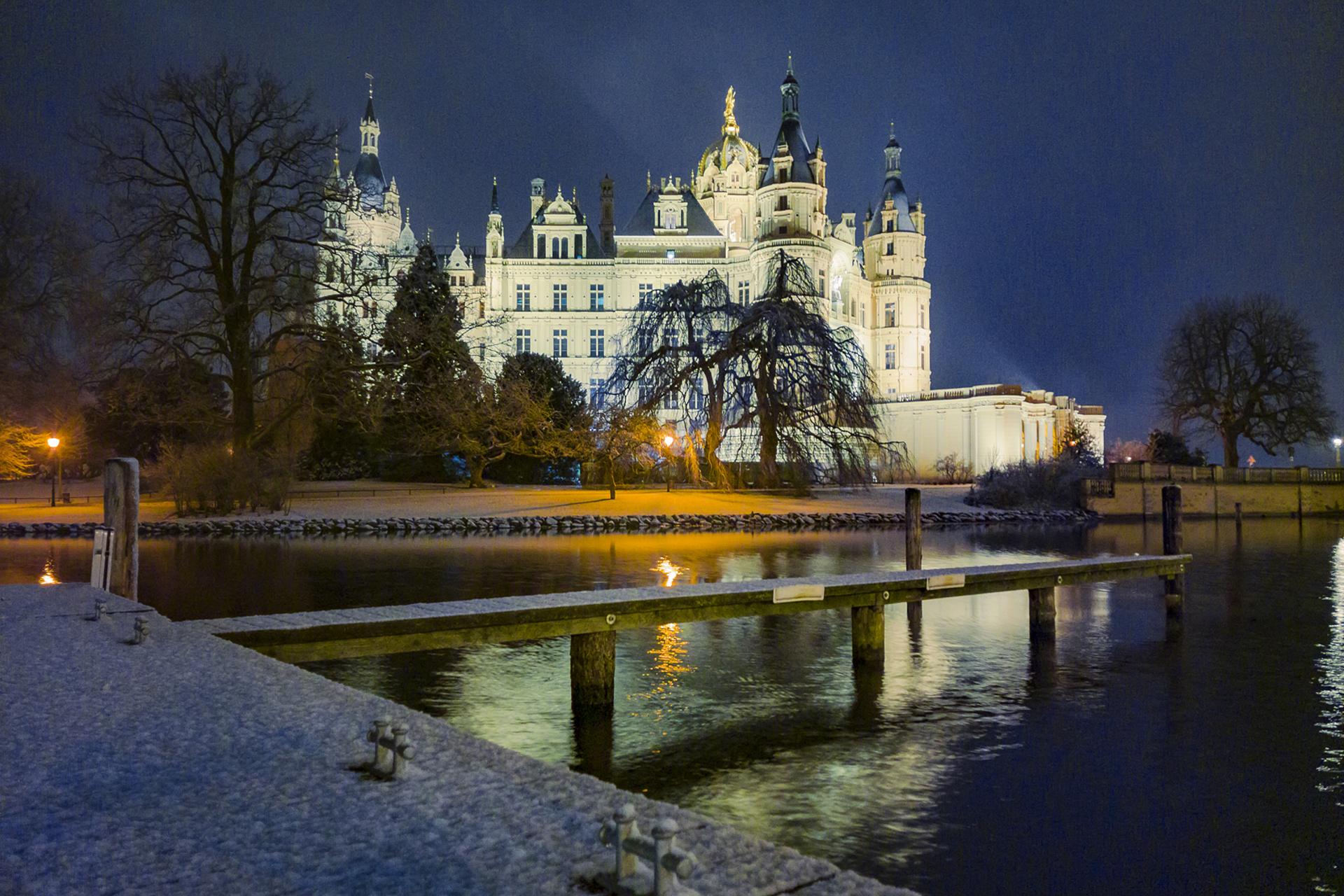 Illuminated historic castle at night reflected in calm water, viewed from a snowy dock under a dark sky.