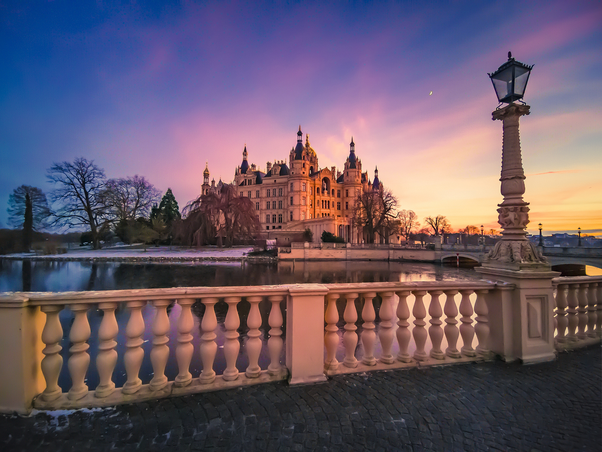 A majestic castle is illuminated by a vibrant sunset sky, viewed from behind an ornate stone railing next to a calm body of water.