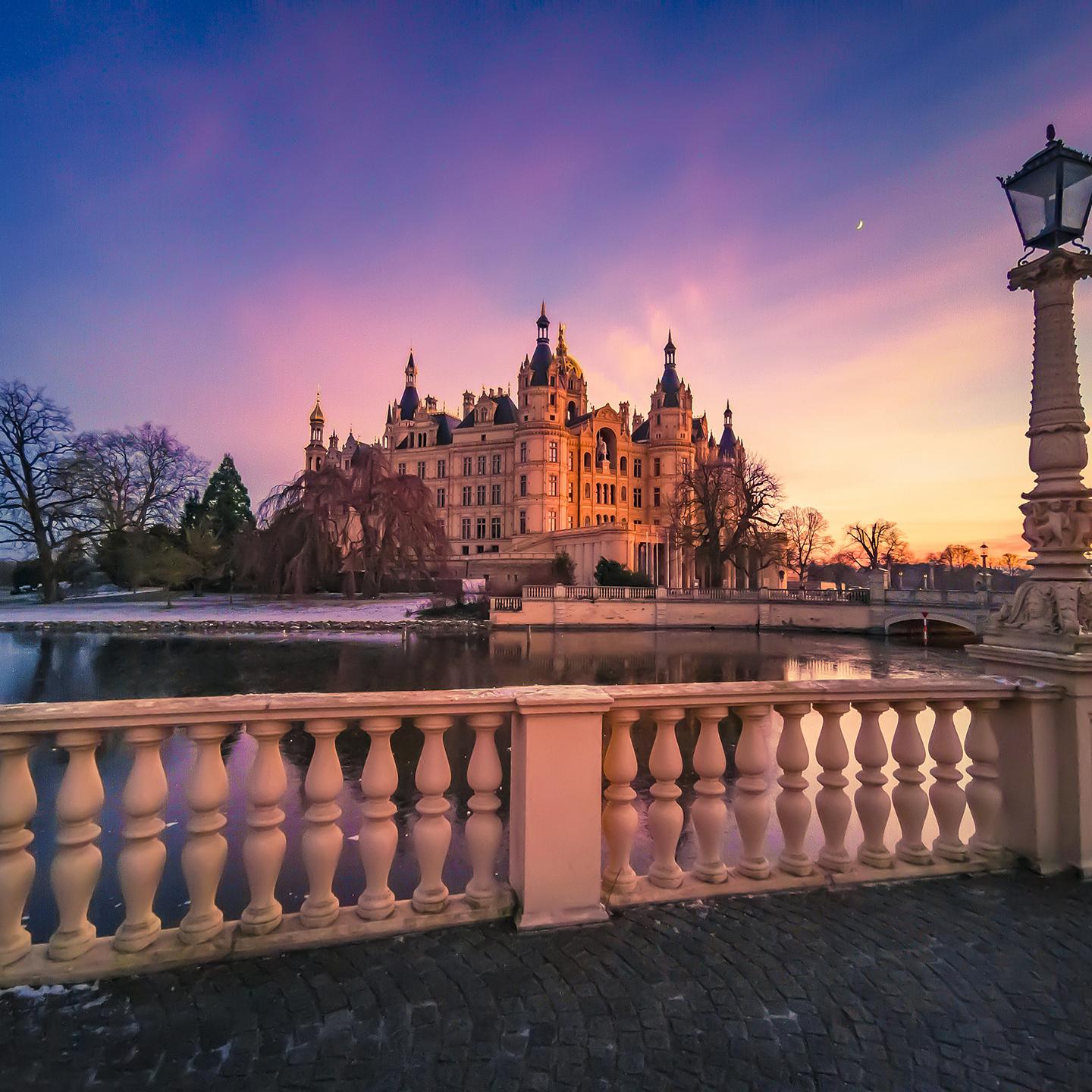 A majestic castle is illuminated by a vibrant sunset sky, viewed from behind an ornate stone railing next to a calm body of water.