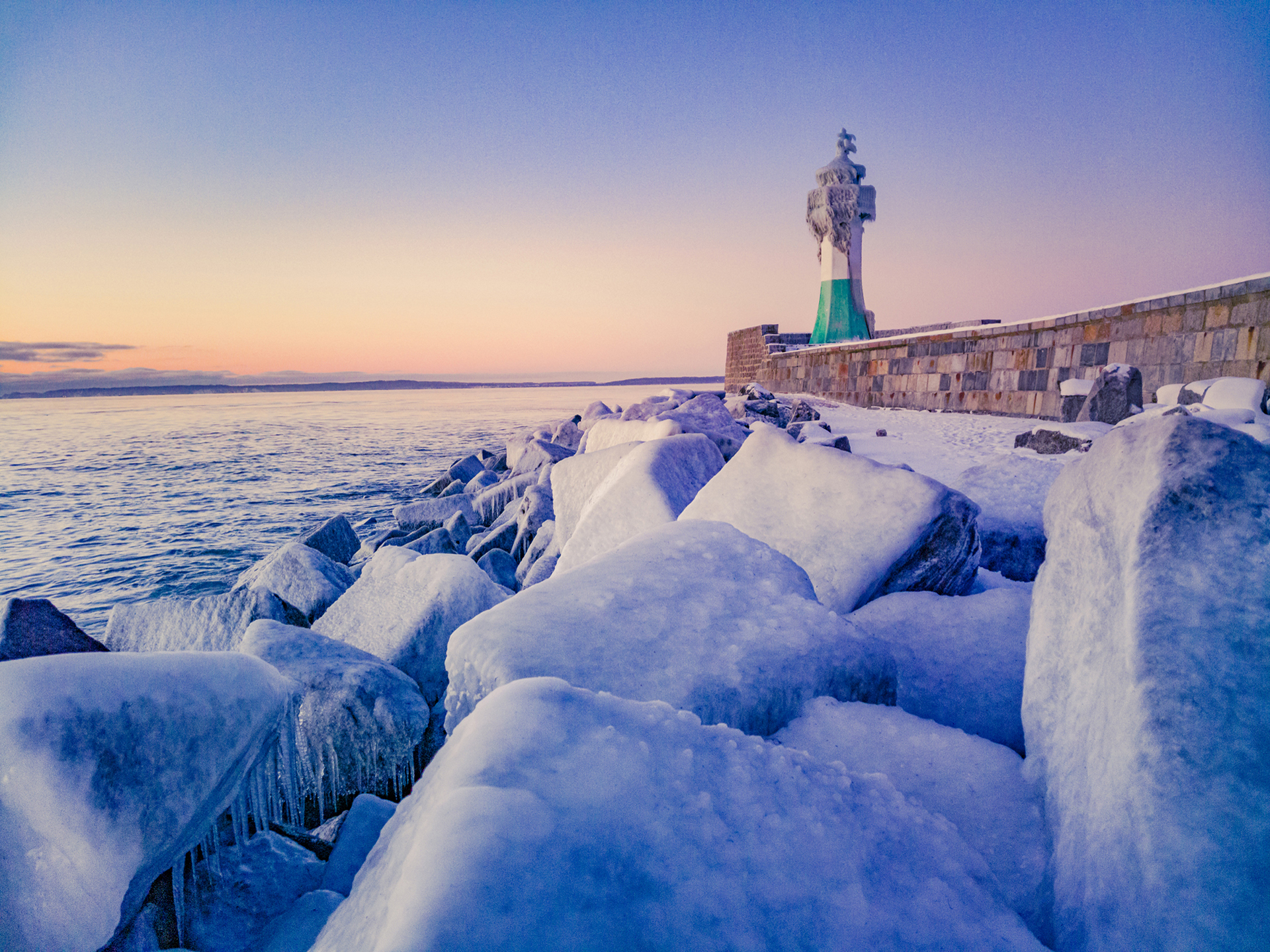 Snow-covered rocks lead to a green and white lighthouse along a frozen shoreline at sunset.