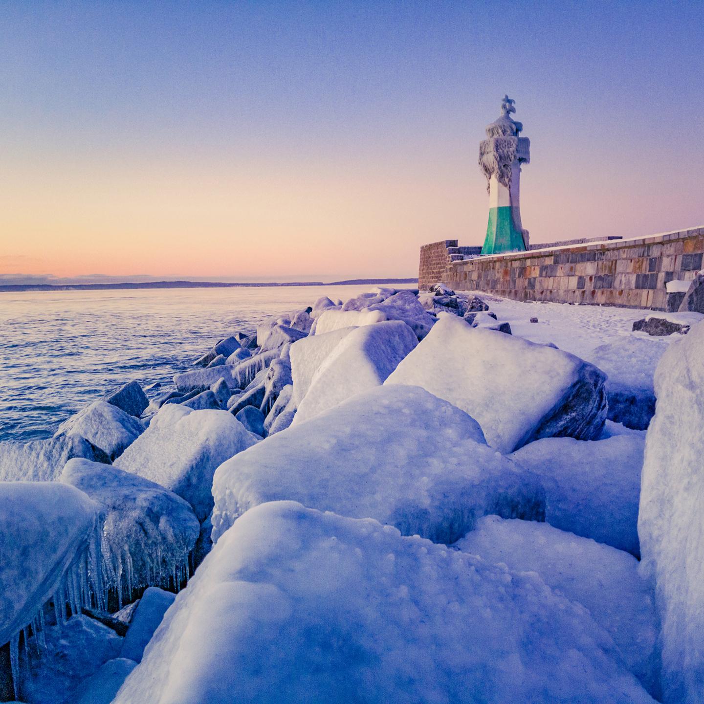 Snow-covered rocks lead to a green and white lighthouse along a frozen shoreline at sunset.