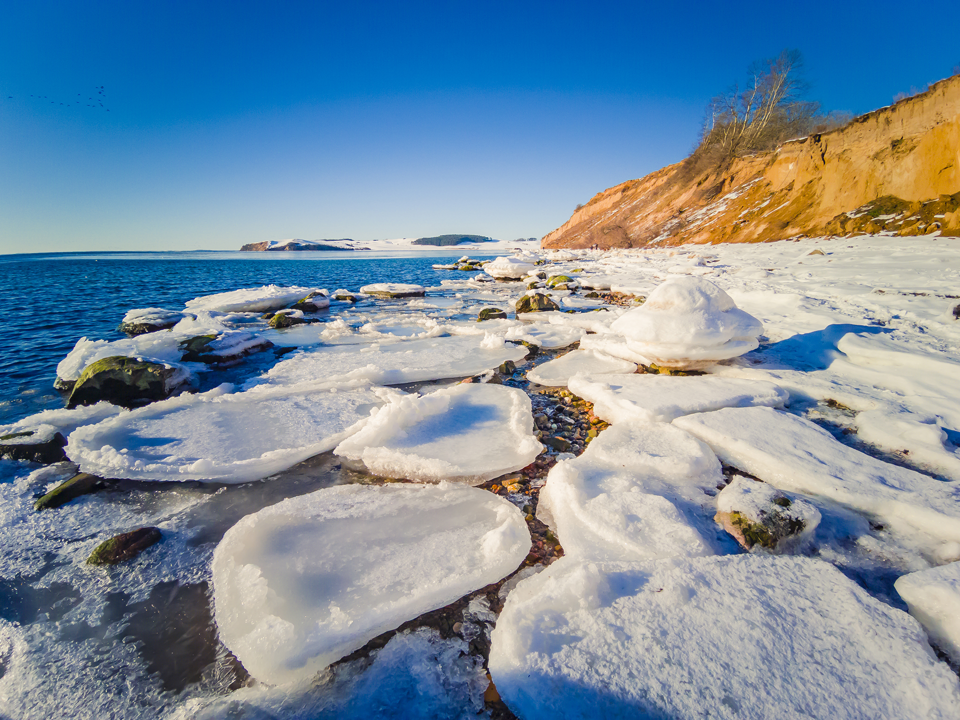 Snow-covered rocks and icy formations line a coastal shoreline beneath a clear blue sky.