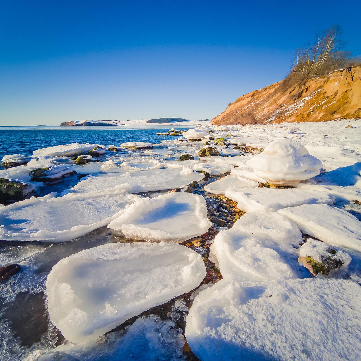 Snow-covered rocks and icy formations line a coastal shoreline beneath a clear blue sky.