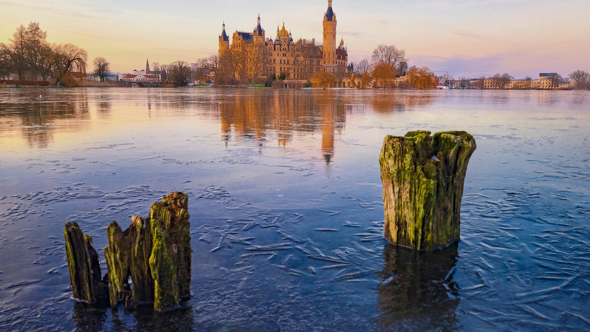 A serene view of a historic castle reflected on a frozen lake at sunset, with weathered wooden posts in the foreground, captured in vivid detail with ZEISS optics.