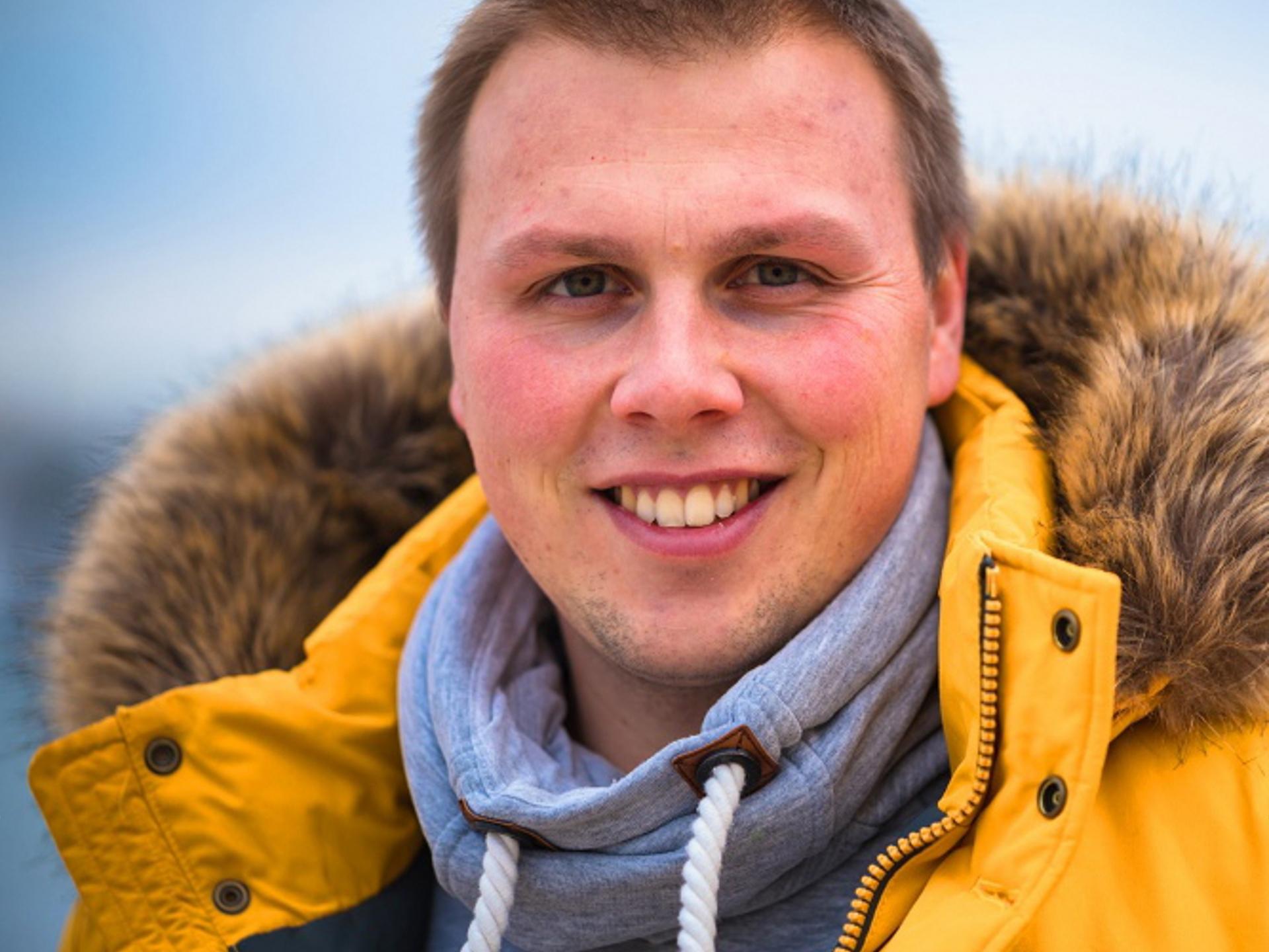 Smiling man wearing a yellow winter jacket with a fur-lined hood and a gray hoodie, standing outdoors.