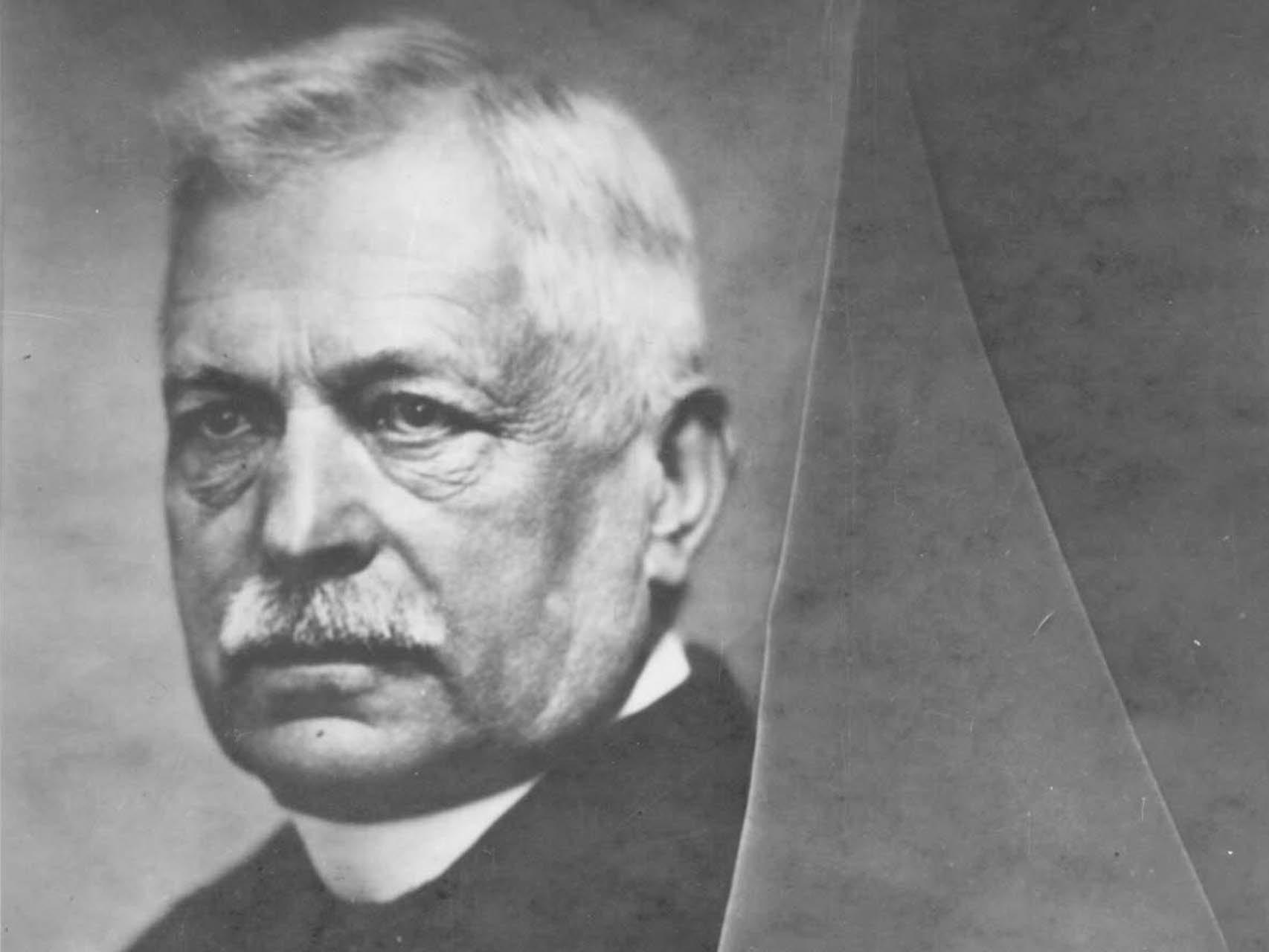 Black and white portrait of an older man with a mustache and short white hair, wearing a suit, with part of the image slightly folded—shown on a ZEISS professional product website.