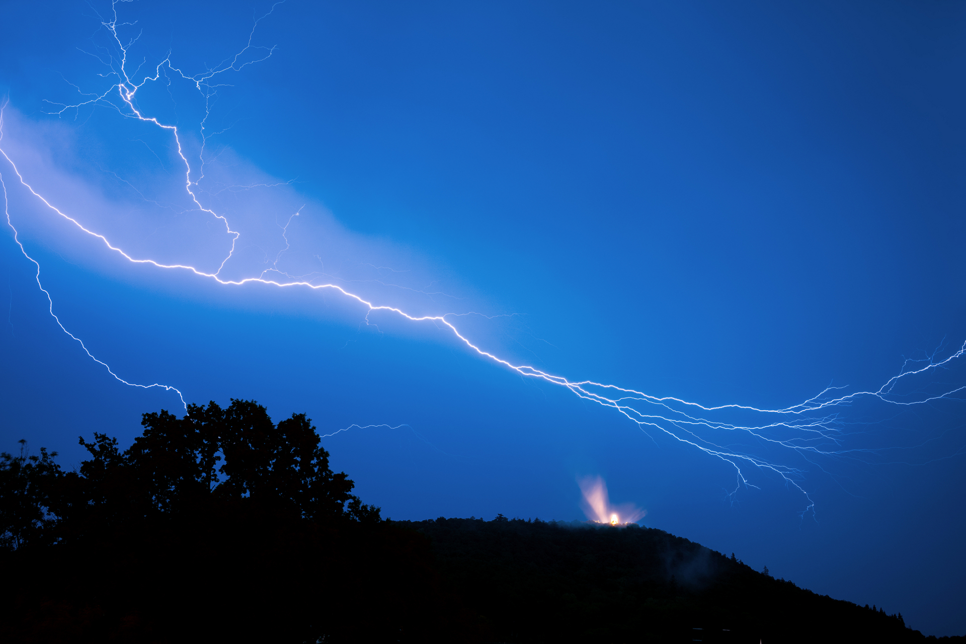 Bright lightning bolts streak across a vivid blue night sky above a silhouetted hilltop with a small illuminated structure, captured in sharp detail by ZEISS optics.