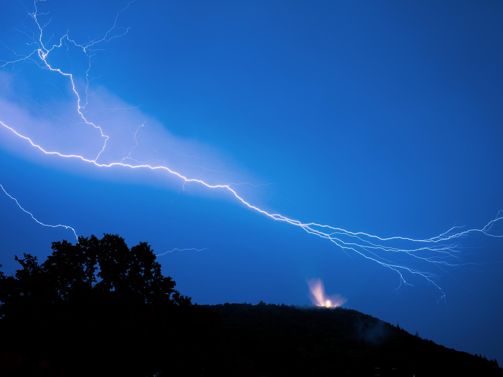 Bright lightning bolts streak across a vivid blue night sky above a silhouetted hilltop with a small illuminated structure, captured in sharp detail by ZEISS optics.