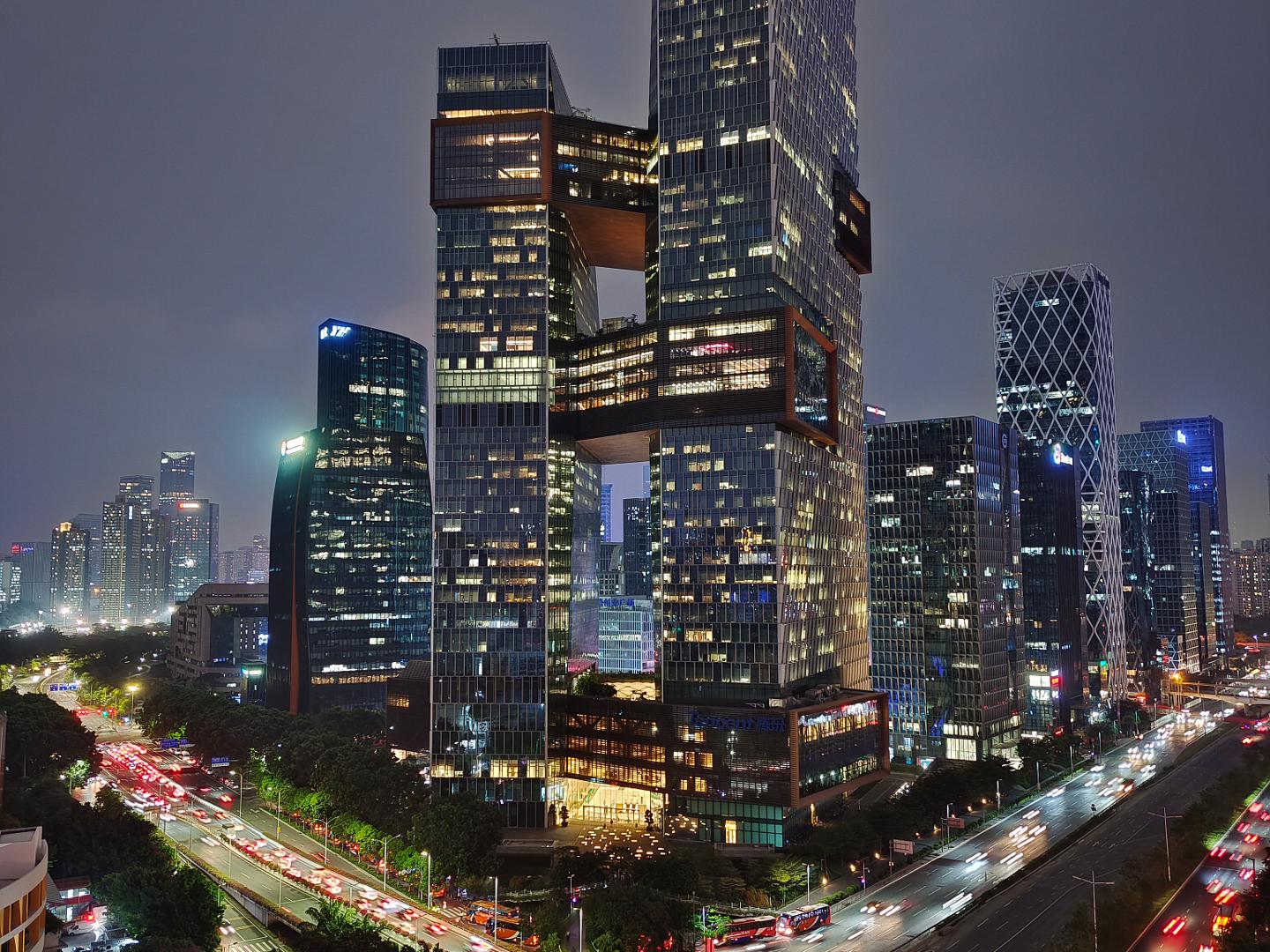 Modern cityscape at night featuring a tall, uniquely structured skyscraper illuminated with lights, surrounded by other high-rise buildings and busy traffic on multi-lane roads.