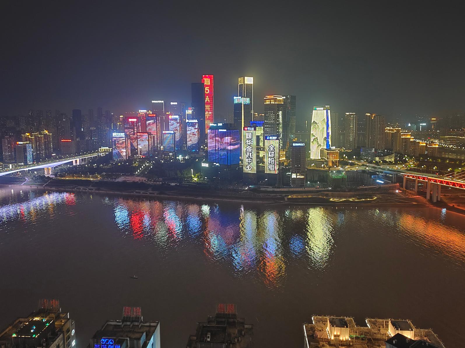 Nighttime cityscape with brightly illuminated skyscrapers reflecting in a wide river, showcasing the clarity and detail captured, ideal for ZEISS optical technology.