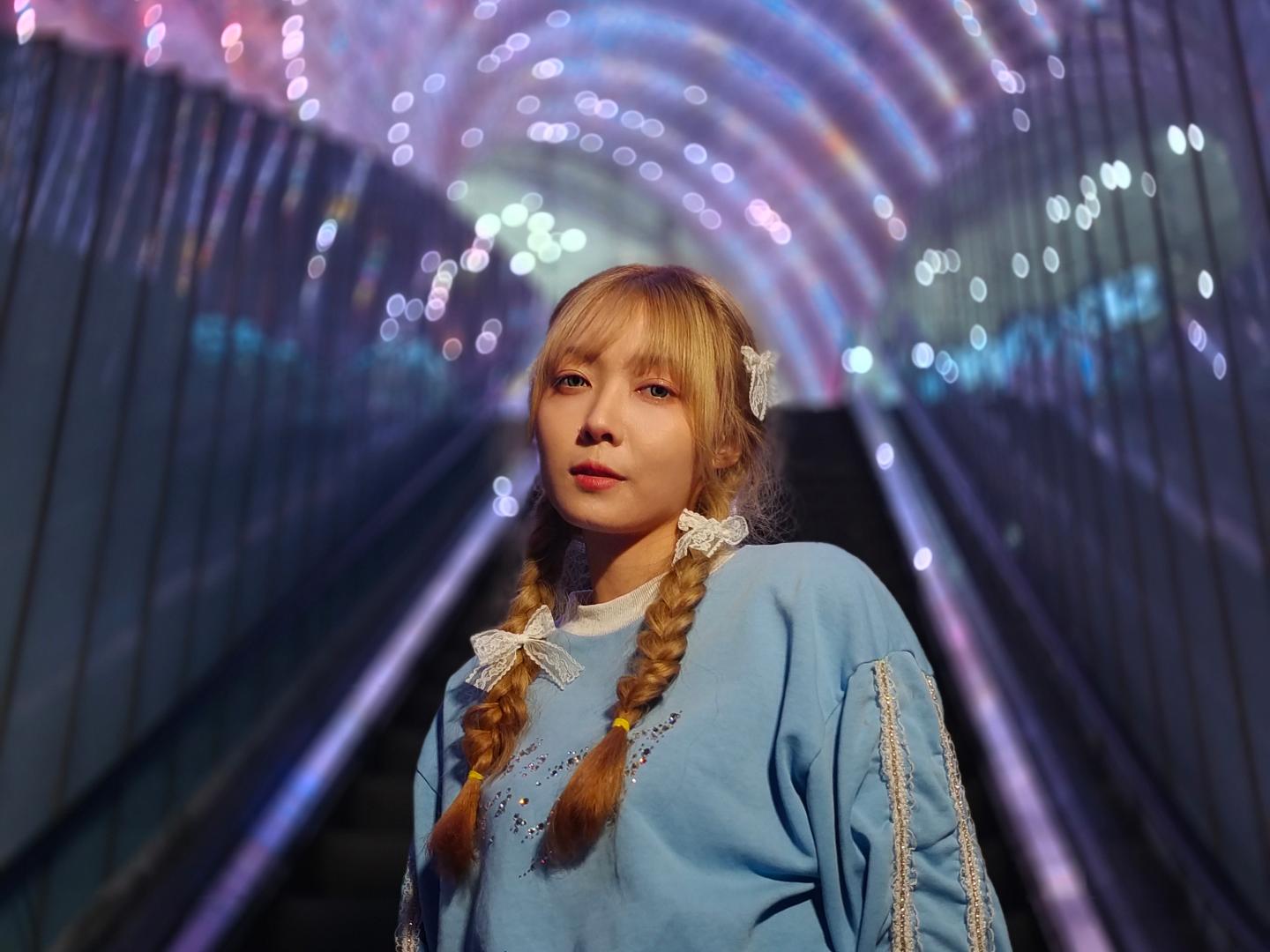 A woman with braided hair stands on an escalator beneath a tunnel of colorful, glowing lights, captured with sharp detail and vibrant colors.