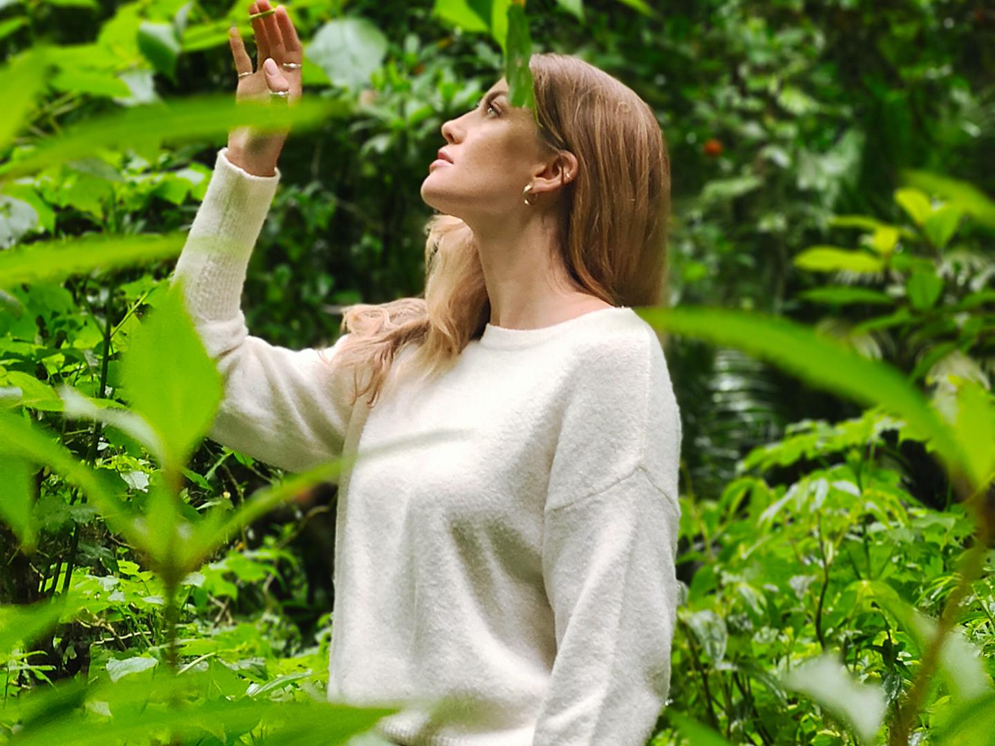 A woman wearing a white sweater stands in a lush green forest, looking upward while surrounded by dense foliage.