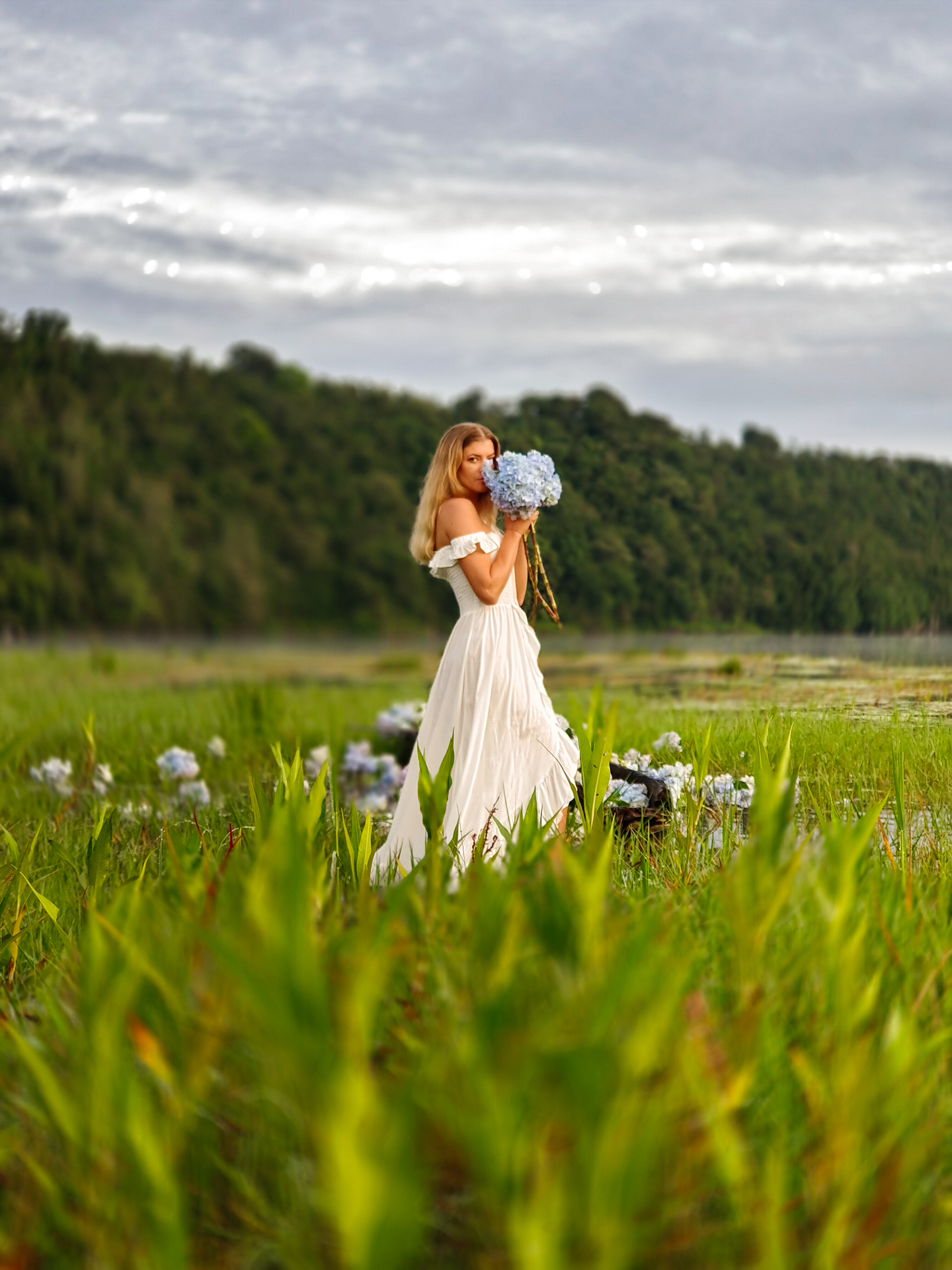 A woman in a flowing white dress stands in a lush green meadow holding a bouquet of blue hydrangeas, with a forest and cloudy sky in the background.