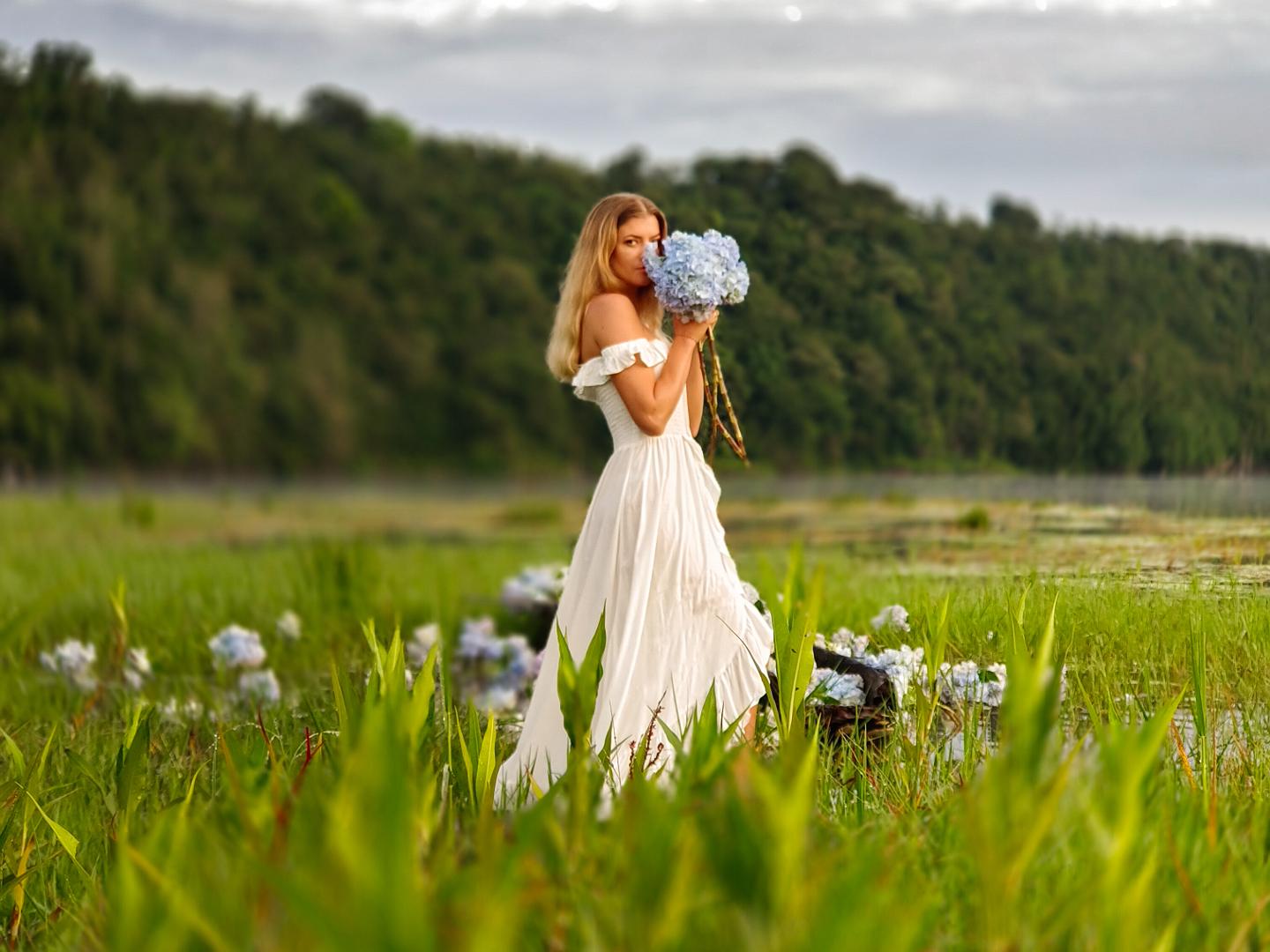 A woman in a flowing white dress stands in a lush green meadow holding a bouquet of blue hydrangeas, with a forest and cloudy sky in the background.