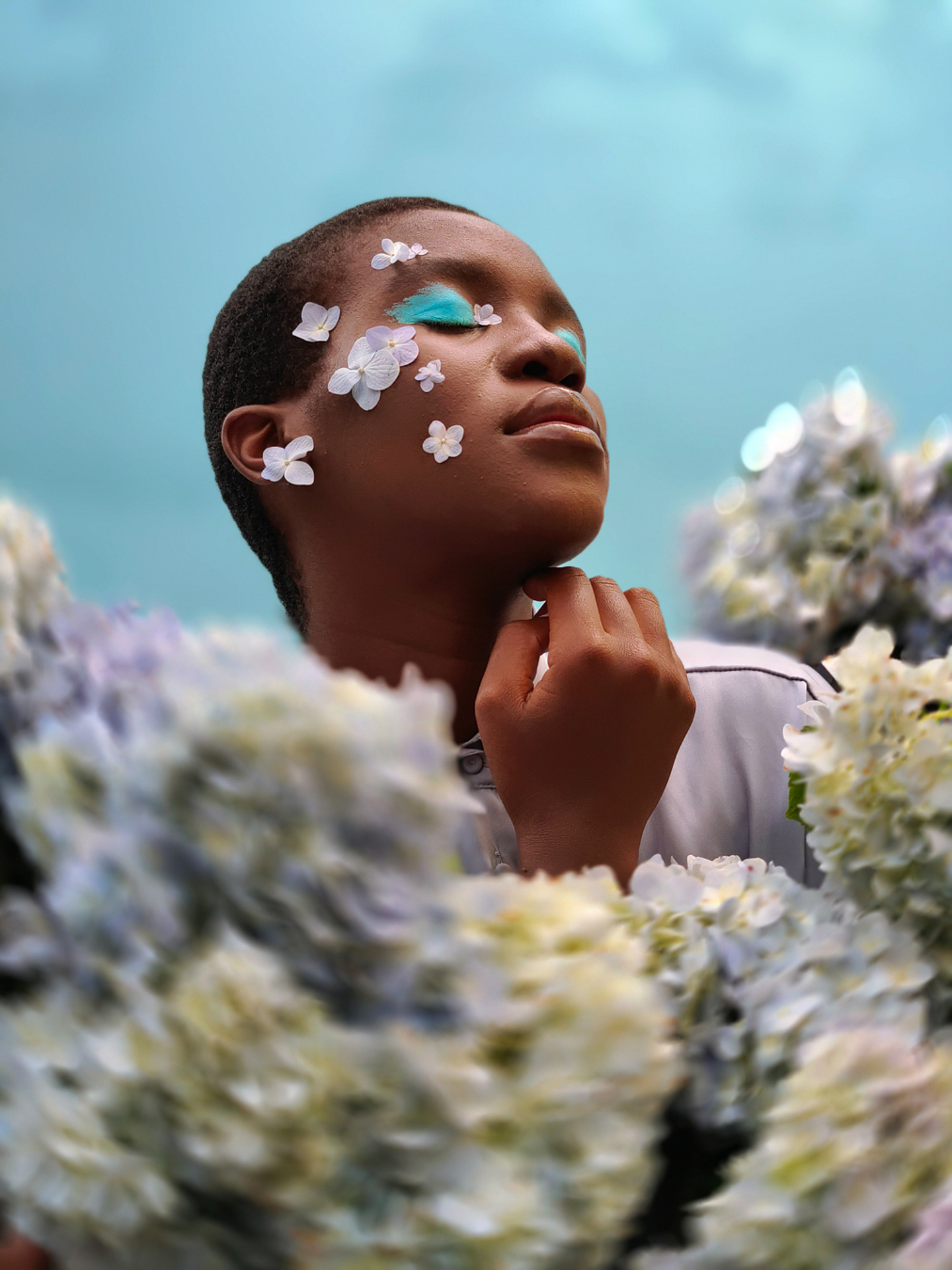 A person with closed eyes and decorative white flower petals on their face poses serenely among clusters of soft-focus hydrangeas under a light blue sky, captured with ZEISS imaging precision.