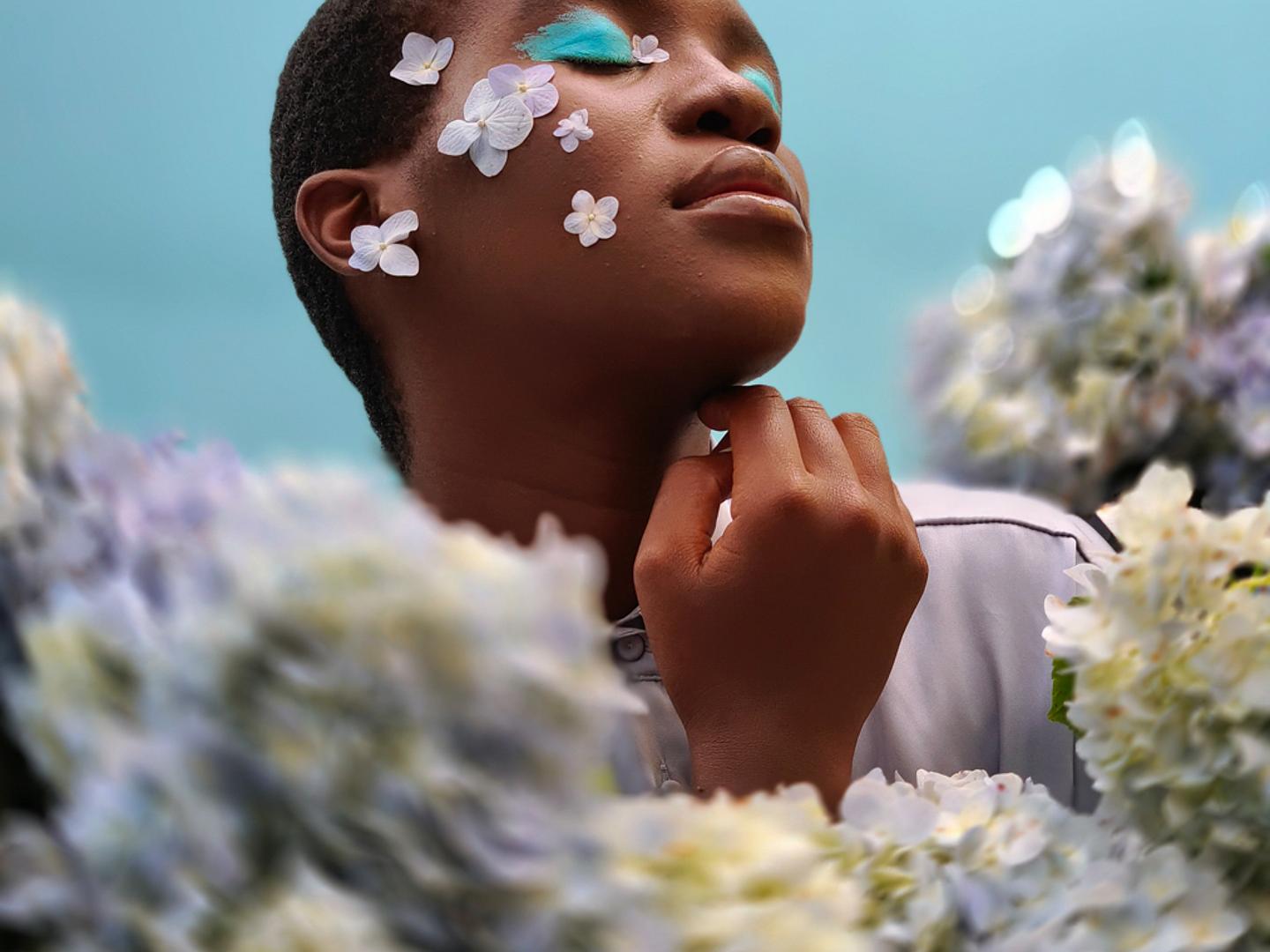 A person with closed eyes and decorative white flower petals on their face poses serenely among clusters of soft-focus hydrangeas under a light blue sky, captured with ZEISS imaging precision.
