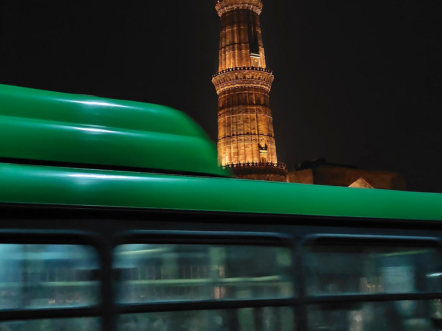 A brightly illuminated historical tower at night stands behind a moving green and yellow bus captured in motion.