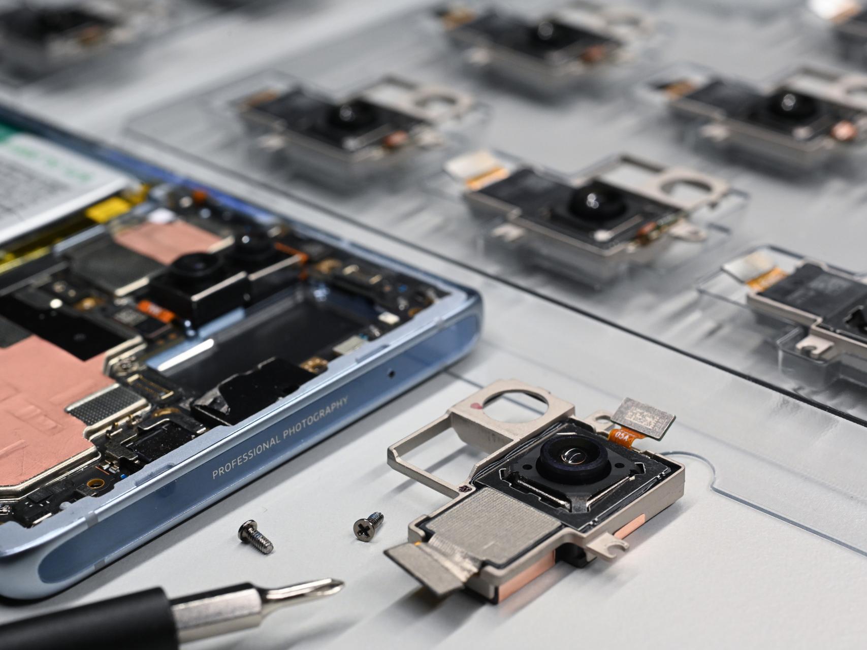 Close-up of disassembled smartphone camera modules, including a ZEISS-branded lens component and internal circuitry, arranged on a workbench.