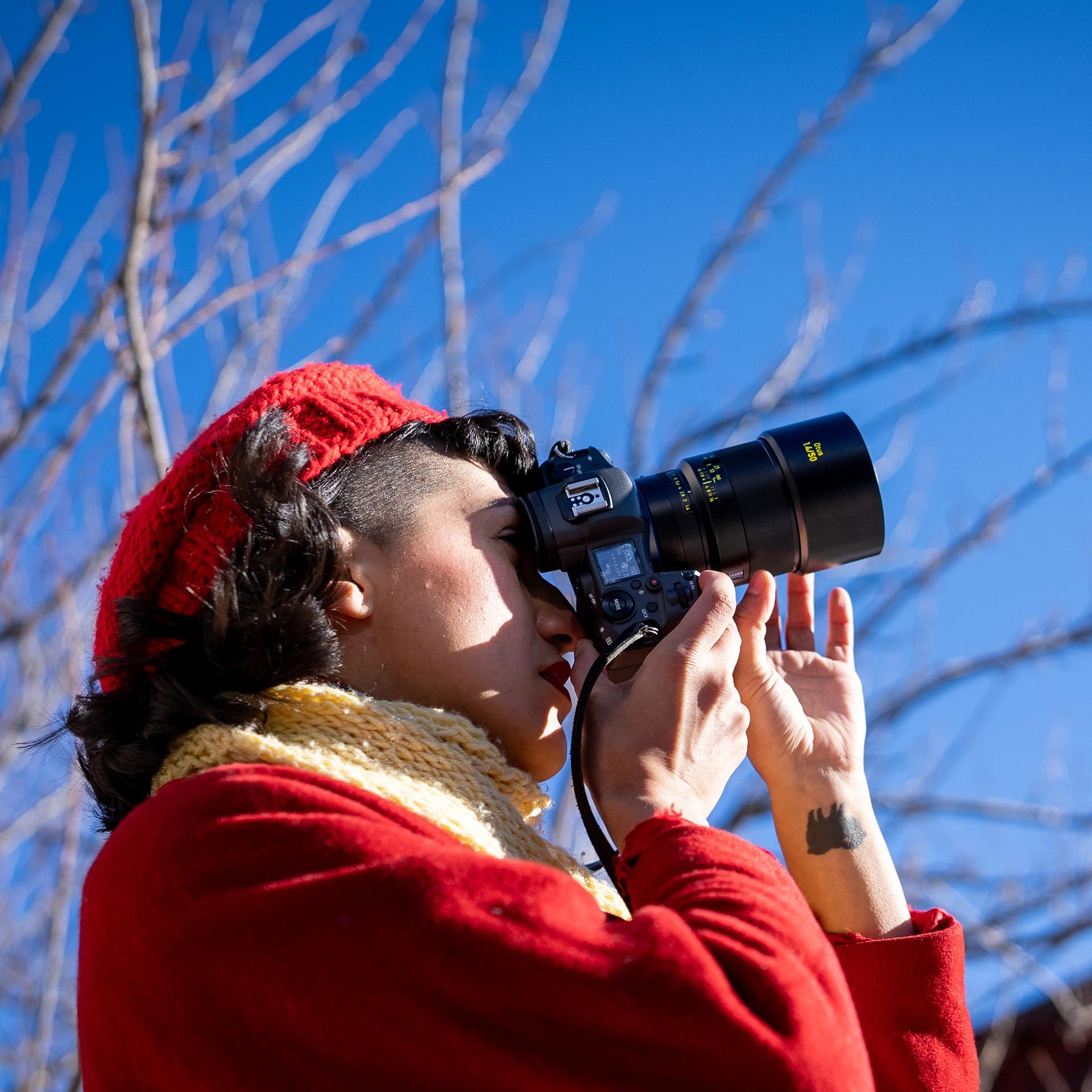 A person wearing a red coat and hat is using a camera equipped with a ZEISS lens outdoors on a clear, sunny day.
