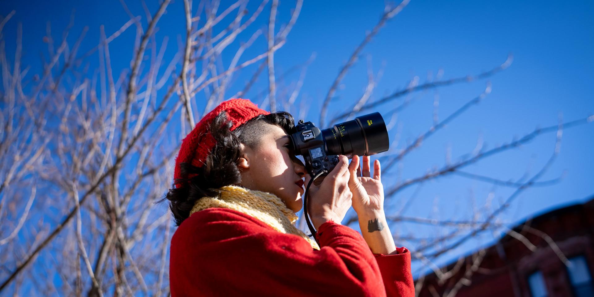 A person wearing a red coat and hat is using a camera equipped with a ZEISS lens outdoors on a clear, sunny day.